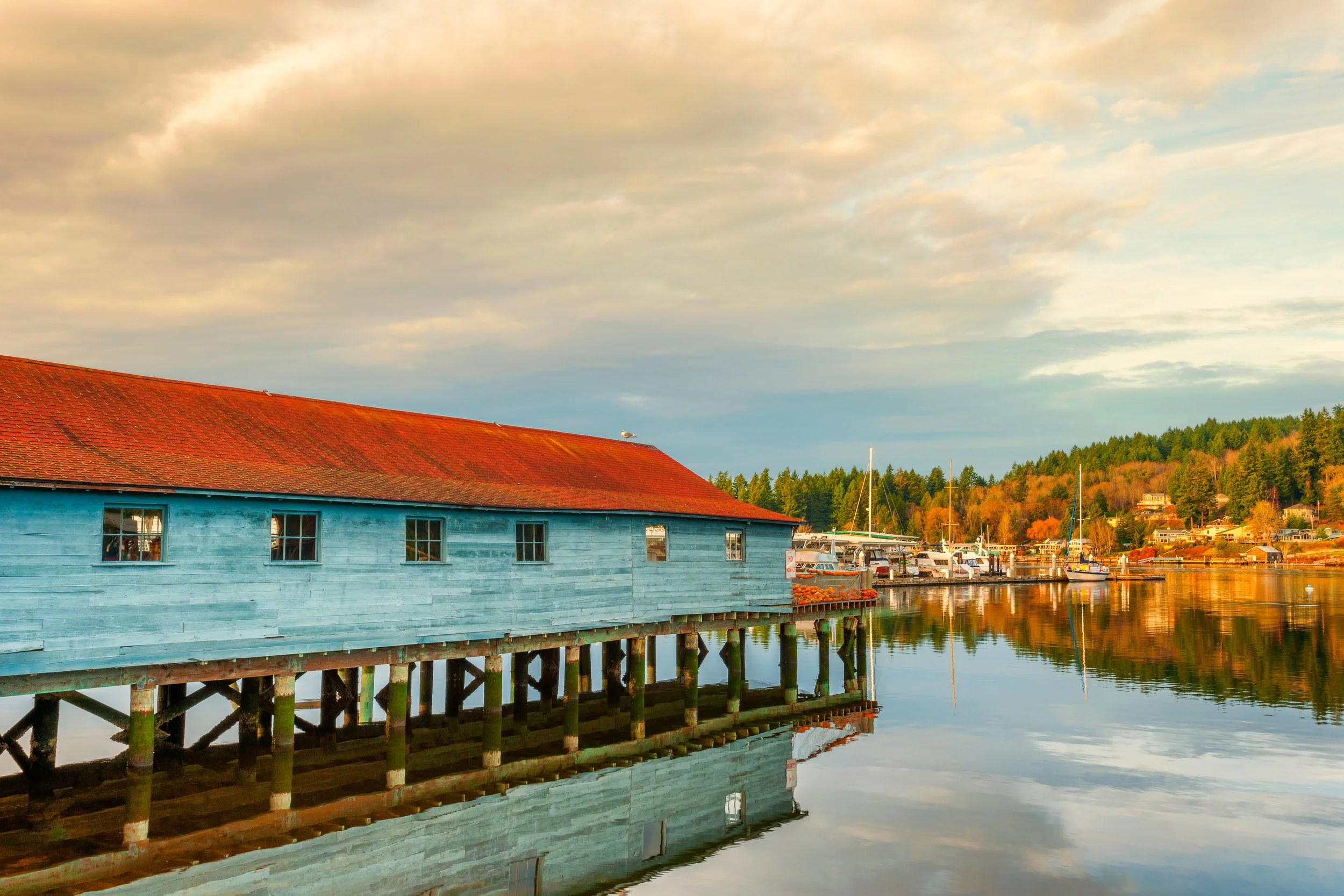A blue wooden building on stilts over calm water, with boats docked along the shoreline and colorful trees in the background under a cloudy sky at sunset.