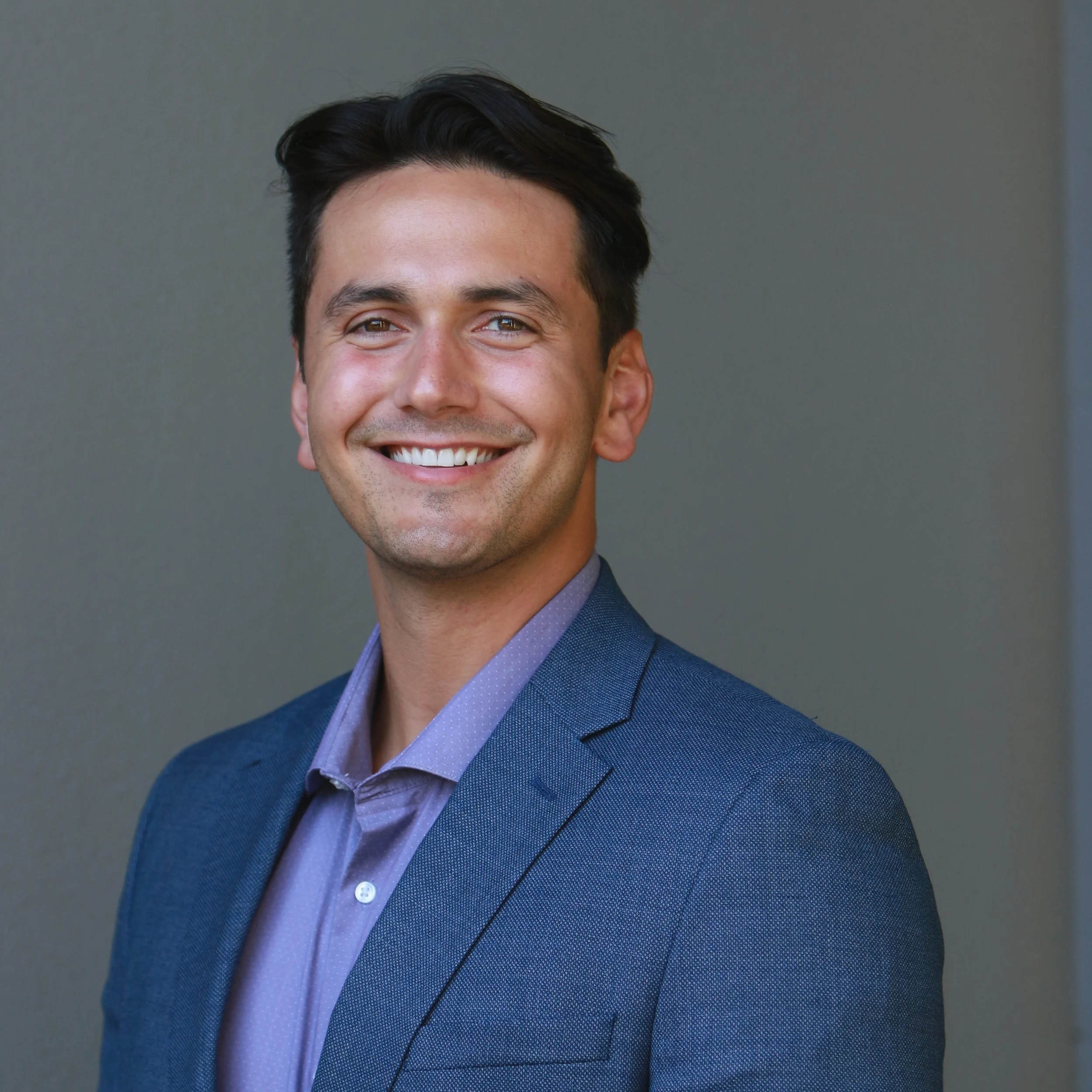 A smiling man with dark hair in a suit and purple dress shirt posing against a plain background.