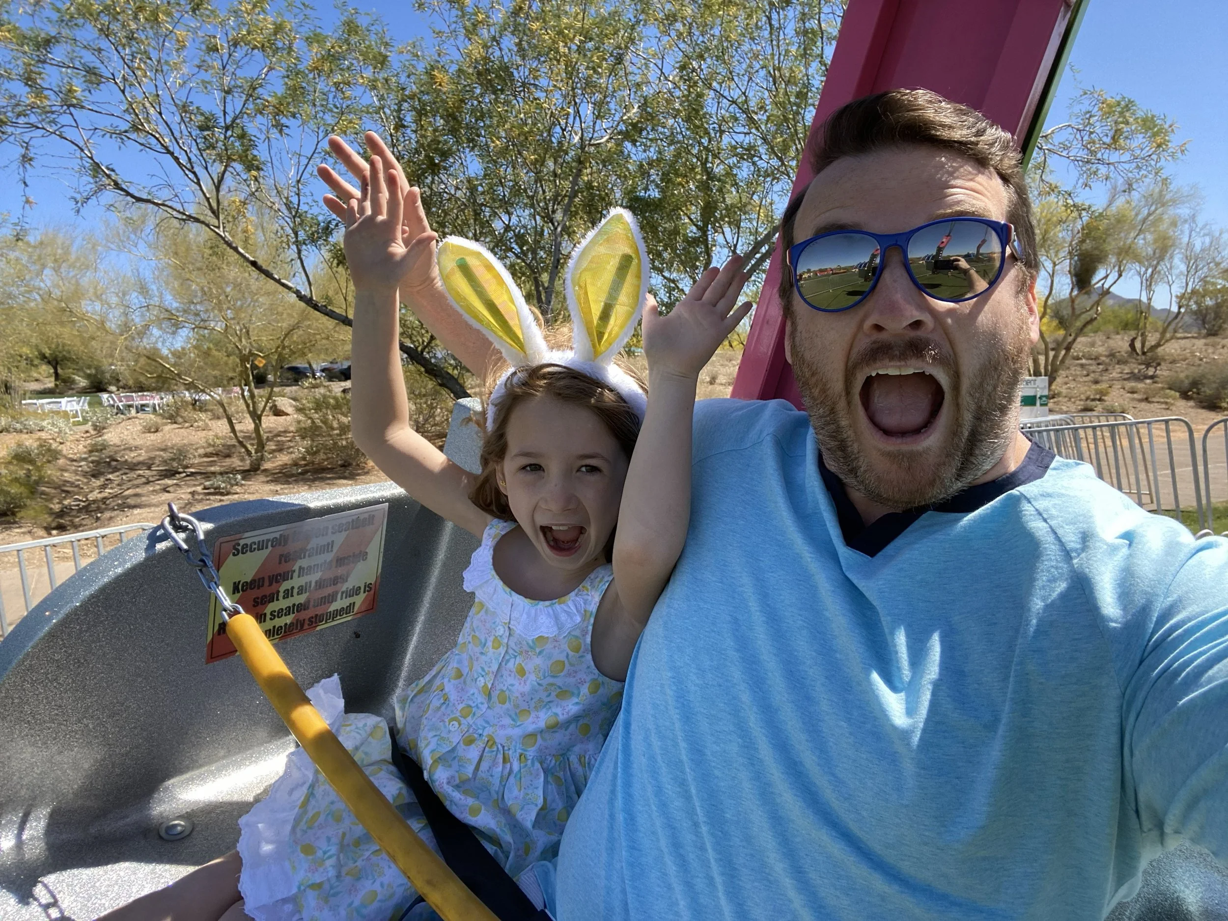 Father and daughter on a roller coaster ride, both with excited expressions; the daughter is wearing bunny ears and a white dress, the father has sunglasses and a beard. The background shows trees and a clear blue sky.