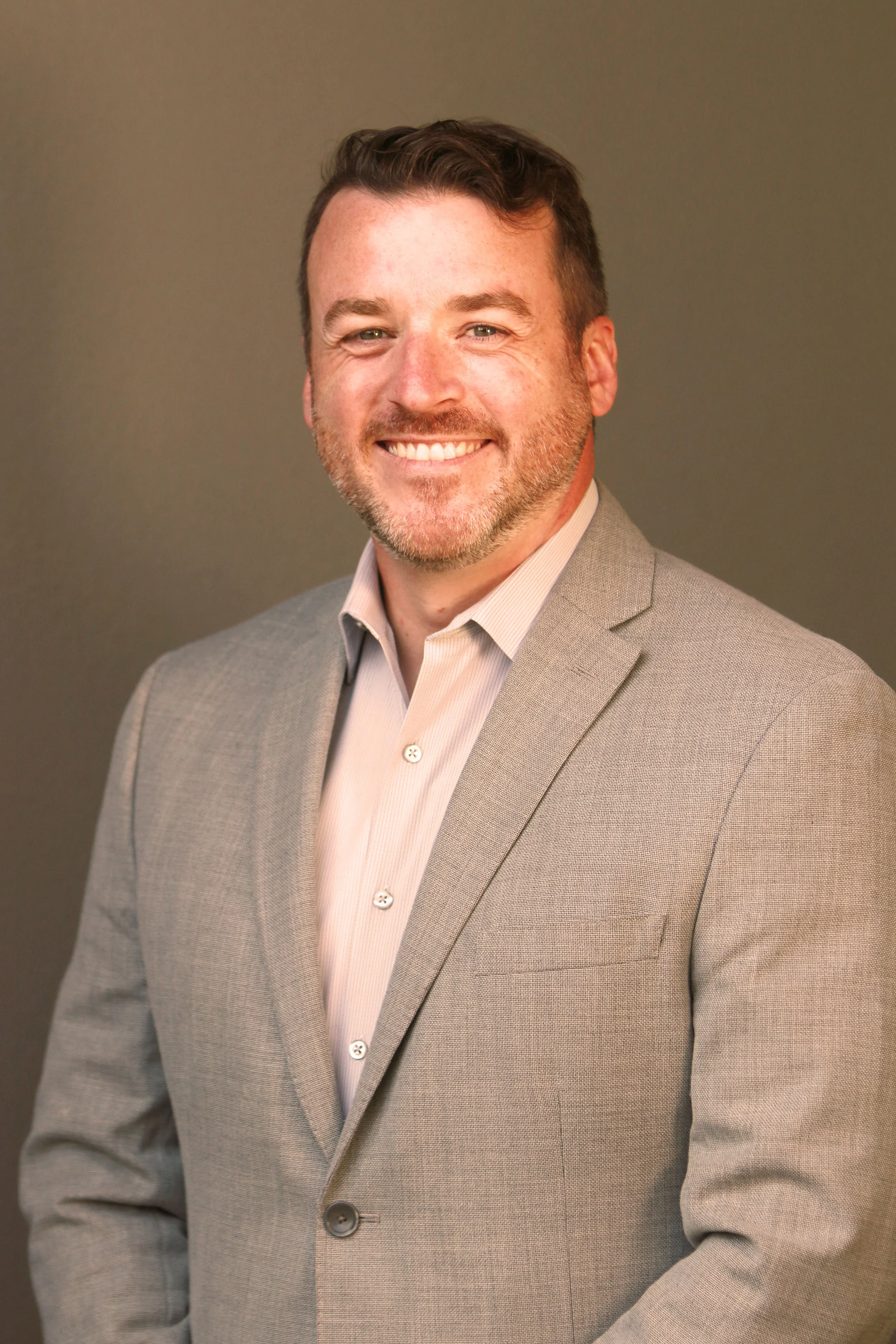 Portrait of a man in a light gray suit jacket and a white collared shirt, smiling against a plain background.