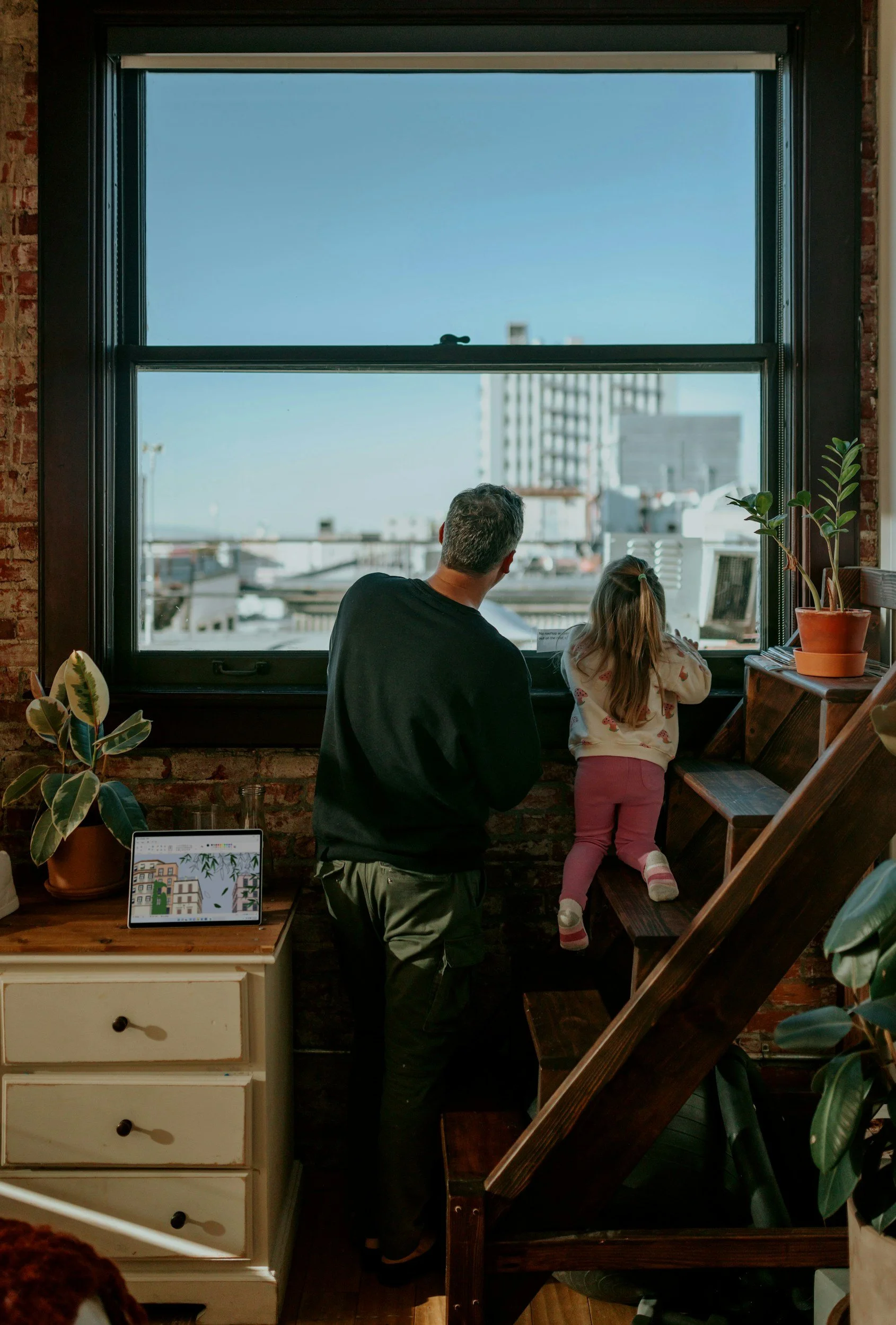 A man and a young girl standing by a large window in a cozy indoor setting with plants, a dresser with drawers, and a tablet on top, watching the cityscape outside.