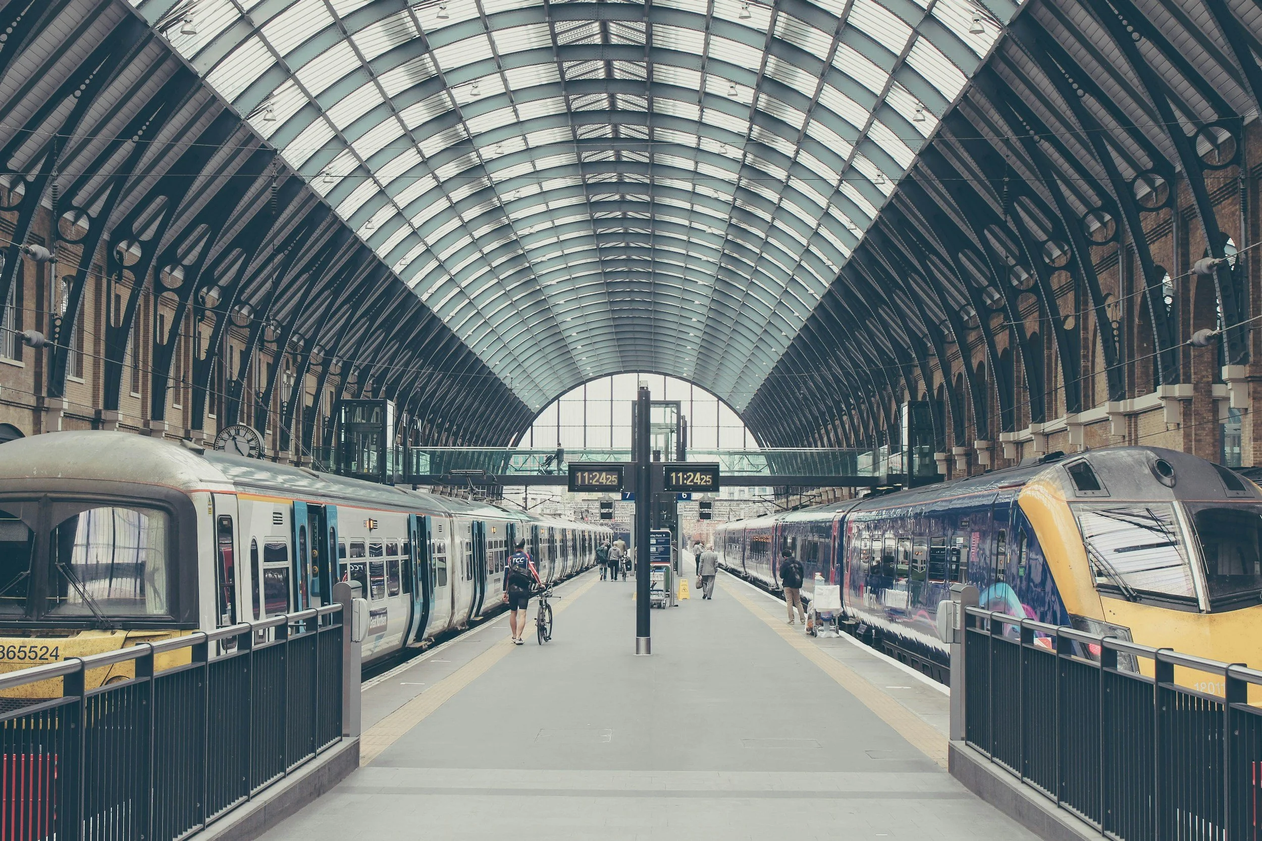 Train station with two trains on either side of the platform under a large arched glass roof, people walking and waiting, digital clocks showing 11:24.