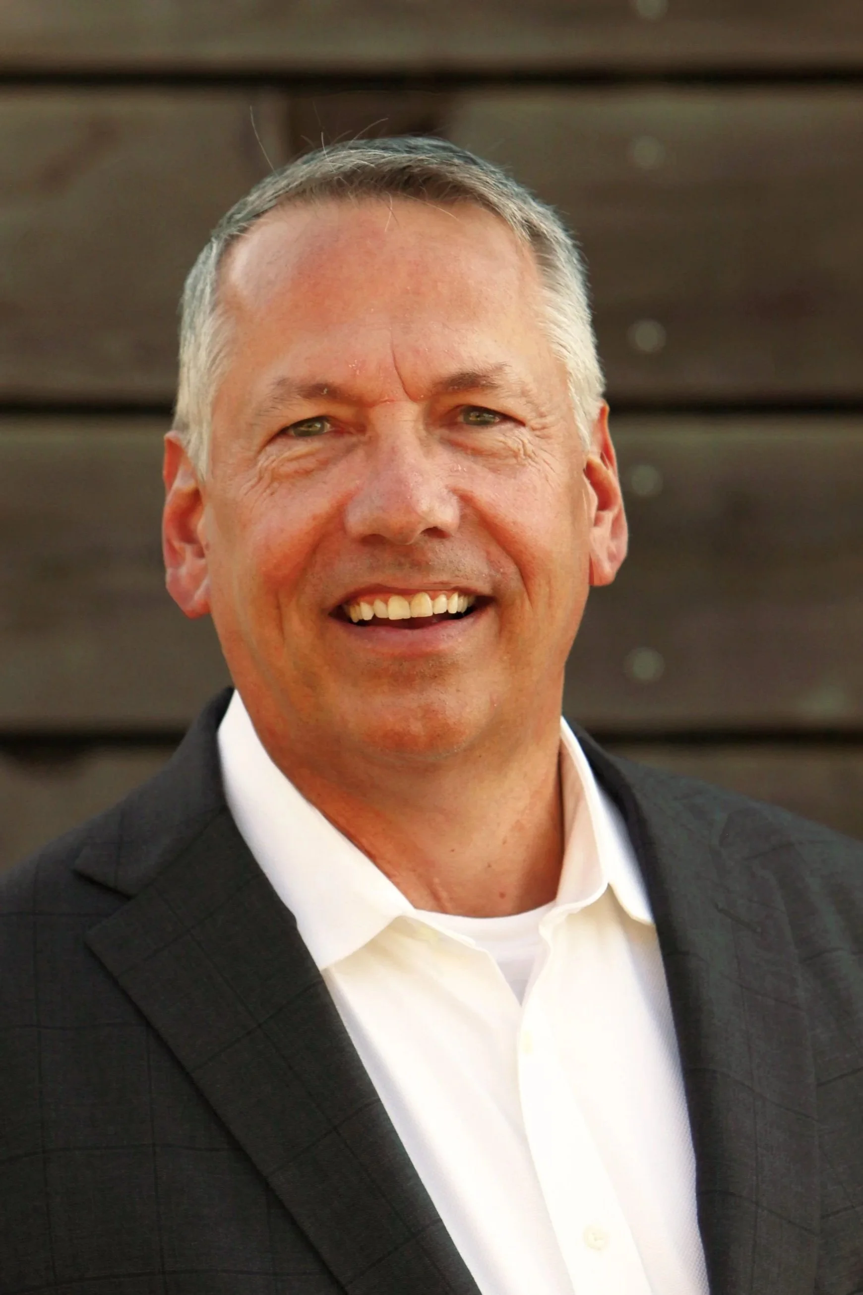 A man smiling, wearing a white shirt and a dark suit jacket, standing in front of a wooden background.