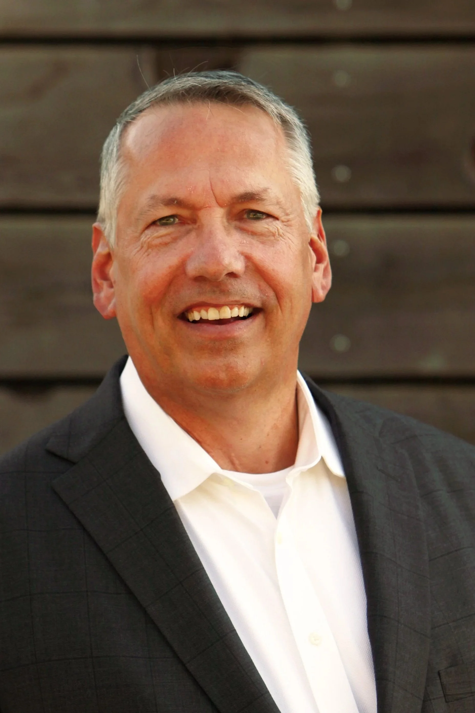 Headshot of a smiling man in a dark suit and white shirt standing outdoors in front of a wooden background.