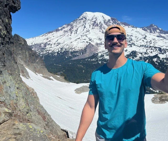 Man taking a selfie in front of snow-capped mountain and rugged terrain with clear blue sky