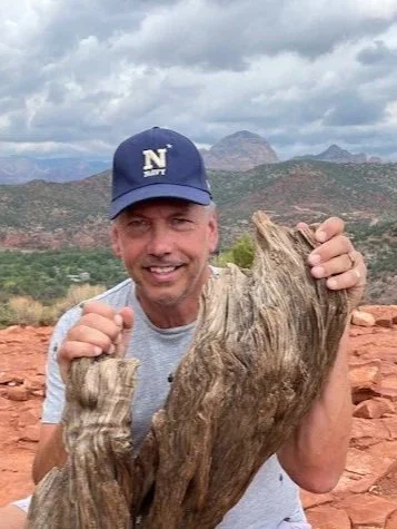 A man smiling and holding a large piece of driftwood outdoors, with a mountainous landscape and cloudy sky in the background.