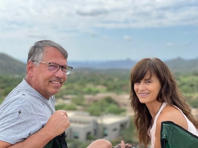 A smiling man and woman sitting outdoors with a scenic landscape in the background