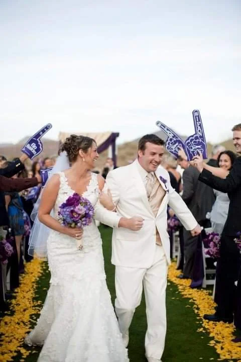 Bride and groom walking down the aisle at their wedding, surrounded by guests holding foam fingers and cheering.