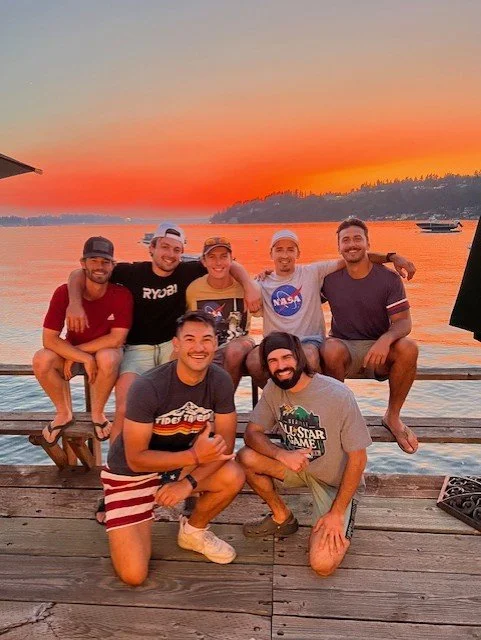 Group of seven young men enjoying a sunset by the water, sitting on a wooden dock with boats in the distance.