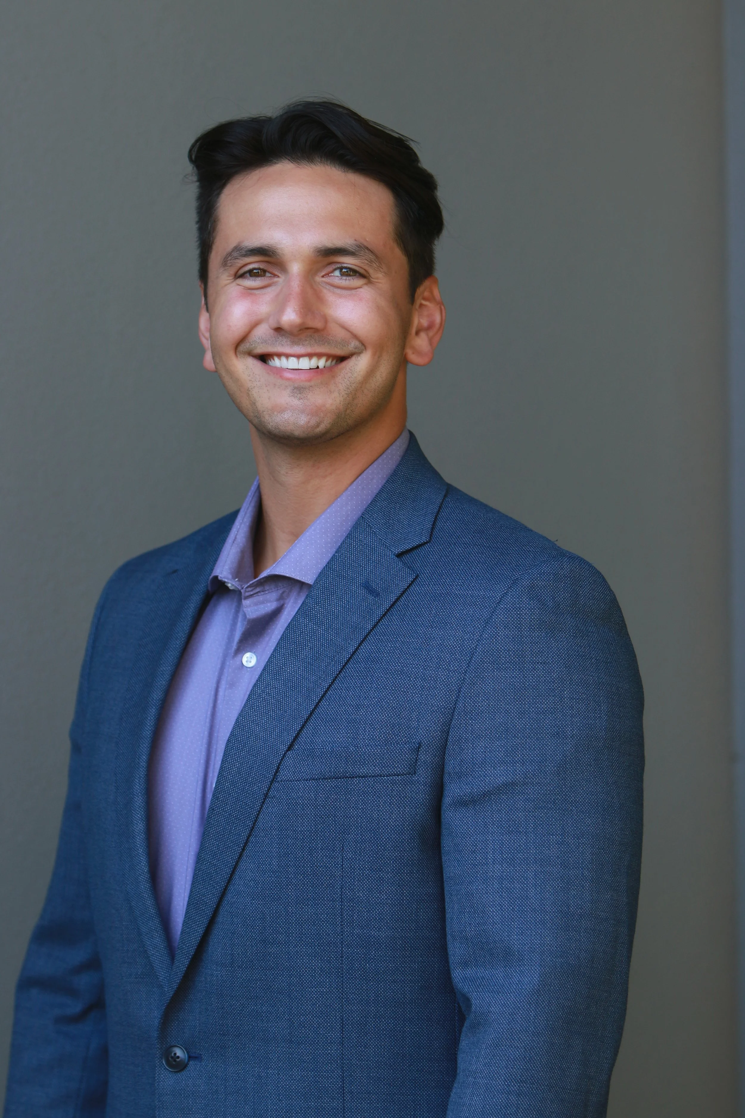 Headshot of a smiling man in a blue suit and purple shirt against a plain background.