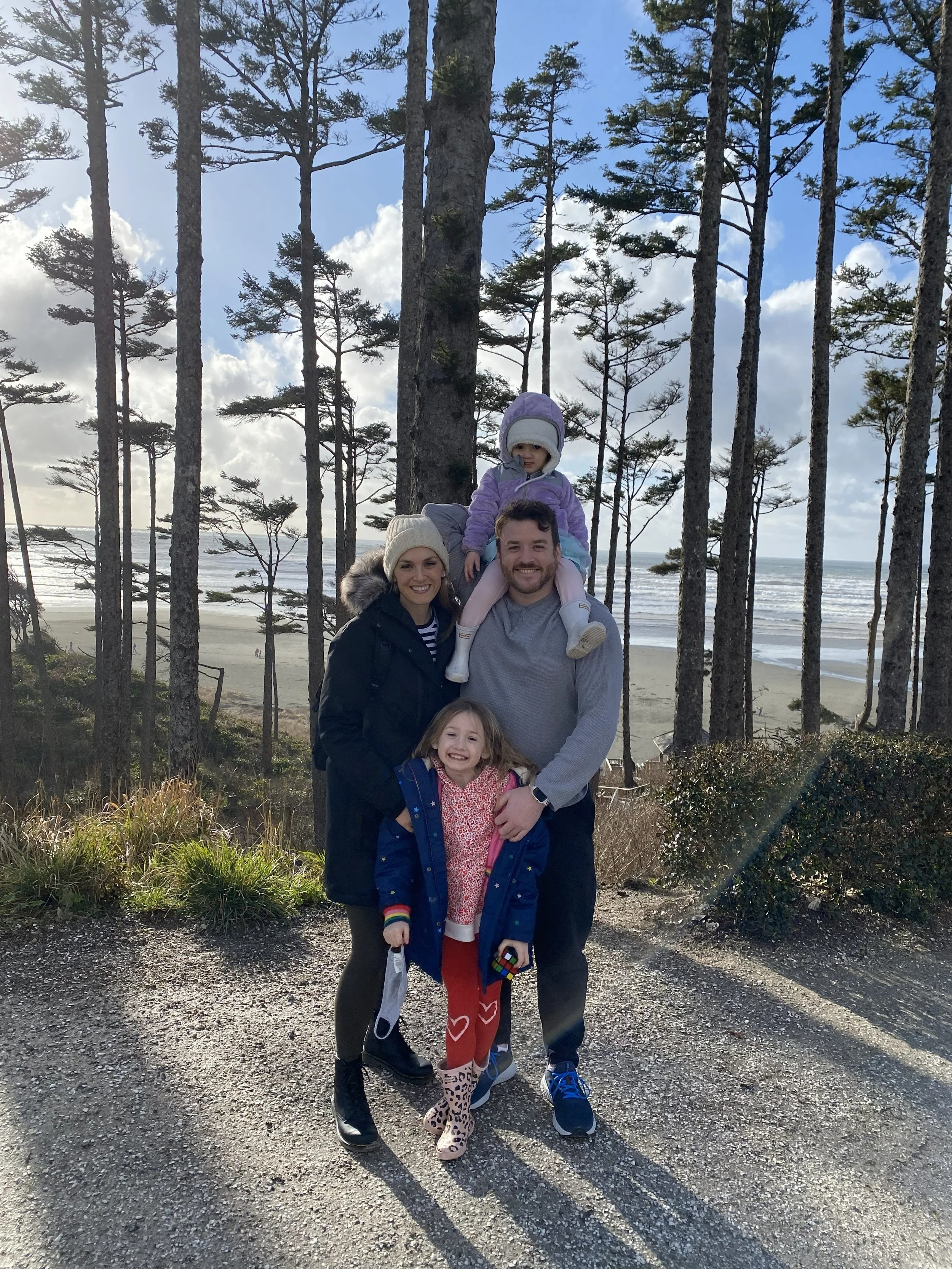 A family of four standing outdoors on a gravel path with tall trees and the ocean in the background. The mother is wearing a coat and beanie, the father is in a gray sweatshirt, and both children are smiling, with one child on the ground and the other sitting on the father's shoulders, all dressed warmly.