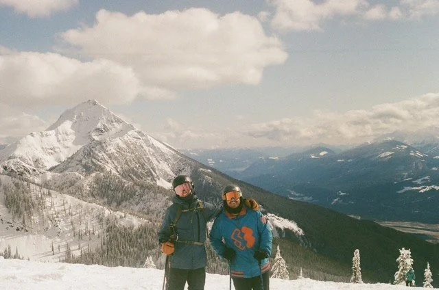 Two people wearing winter gear standing on snow-covered ground with a mountain and cloudy sky in the background.