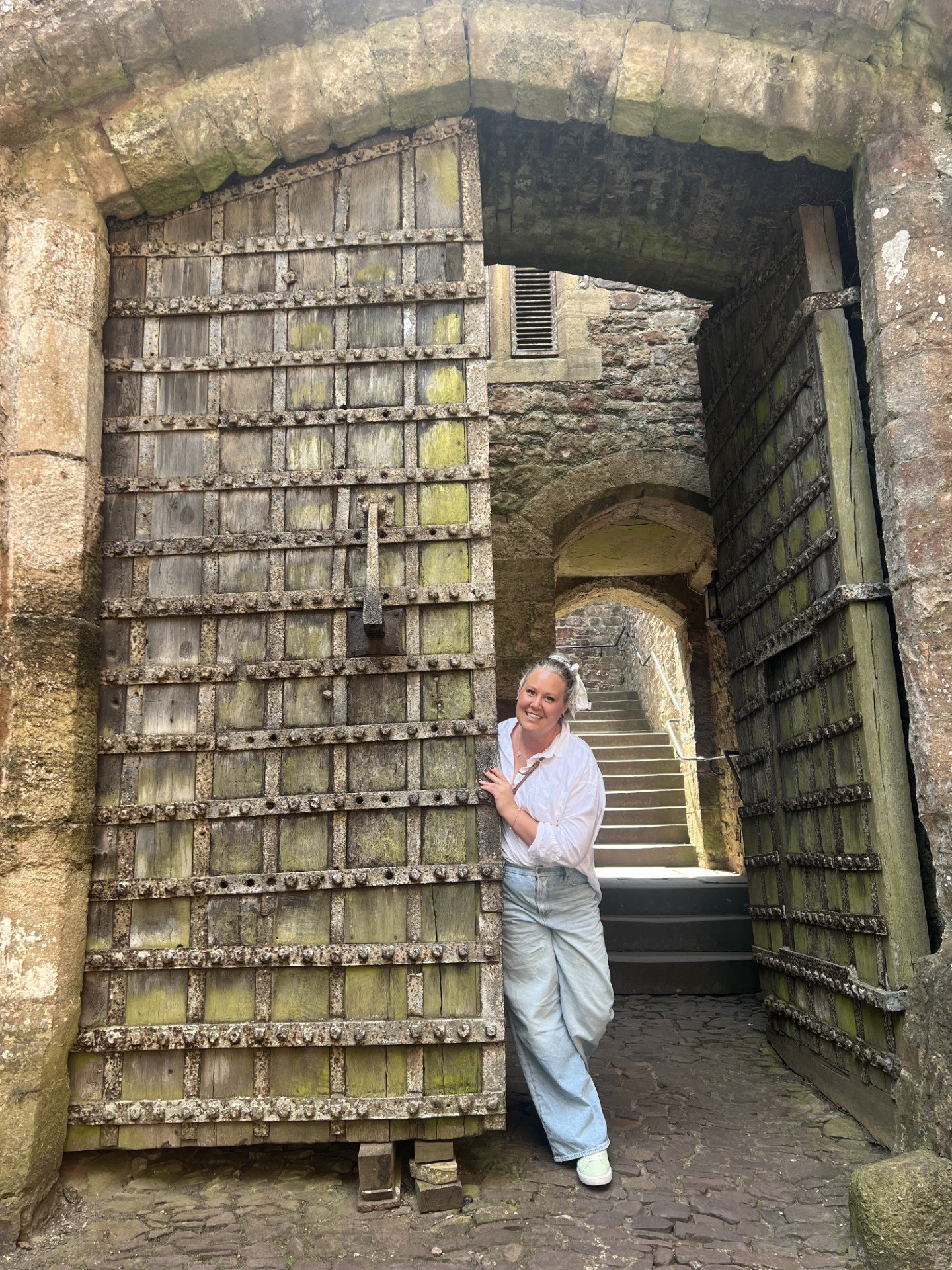 A smiling woman wearing a white shirt and light blue jeans peeking out from behind a large, weathered wooden door in an old stone building. The door is partially open, revealing stone stairs leading up in the background.