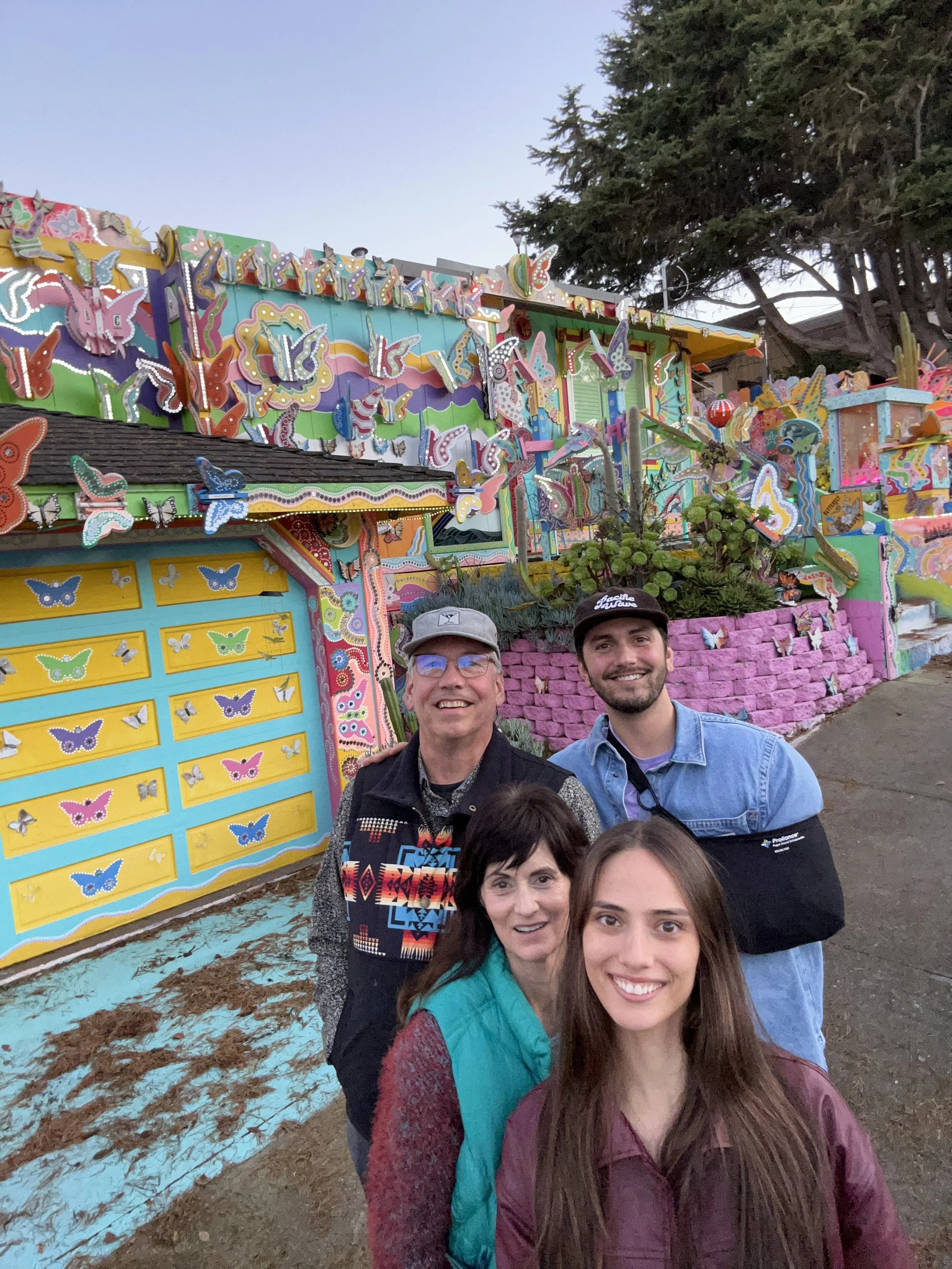 Four people smiling for a photo outside a colorful, butterfly-themed art installation with pink and purple brick accents, trees in the background, and a partly cloudy sky.