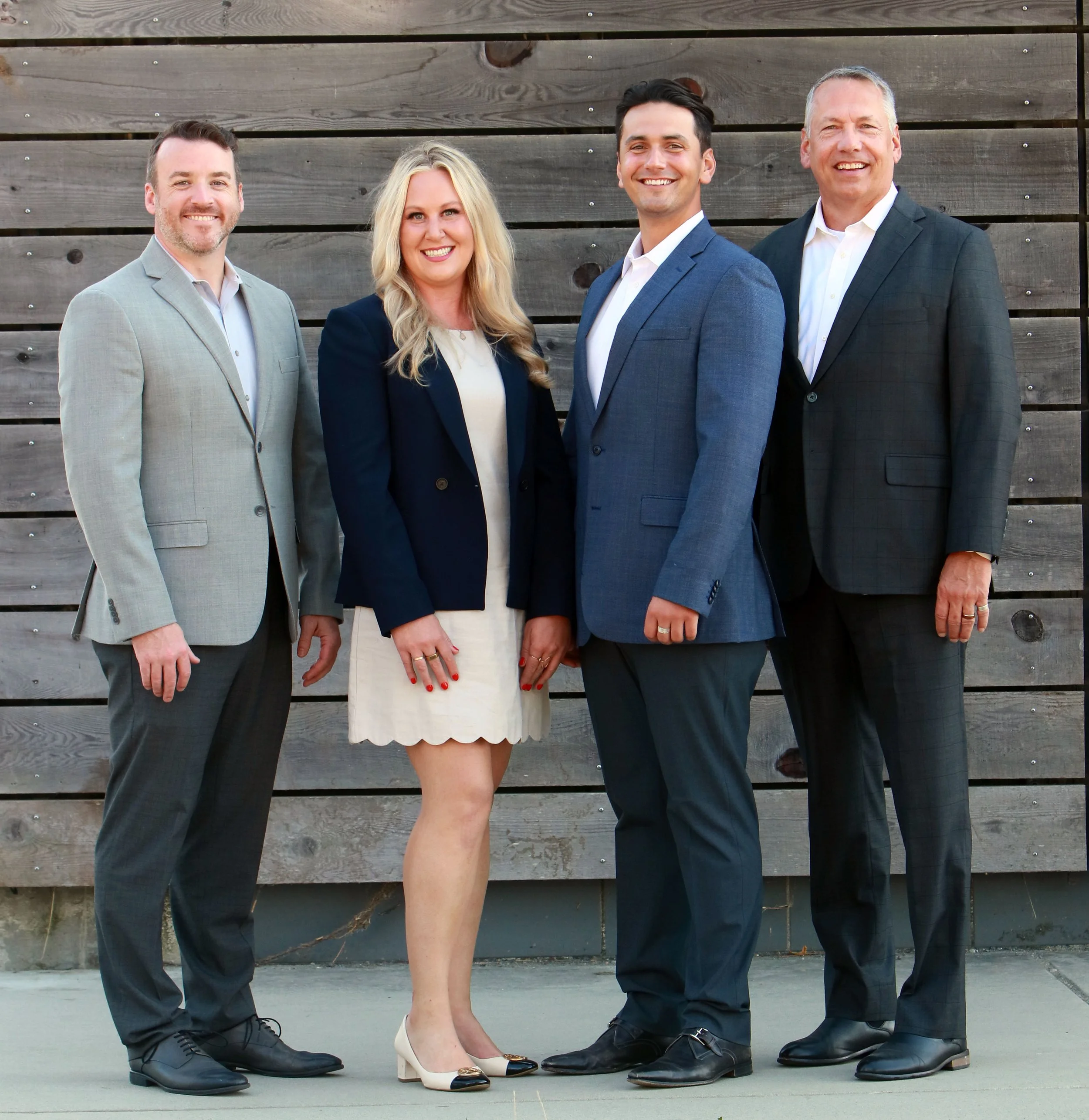Group of four professionally dressed people standing outdoors in front of a wooden wall, smiling at the camera.