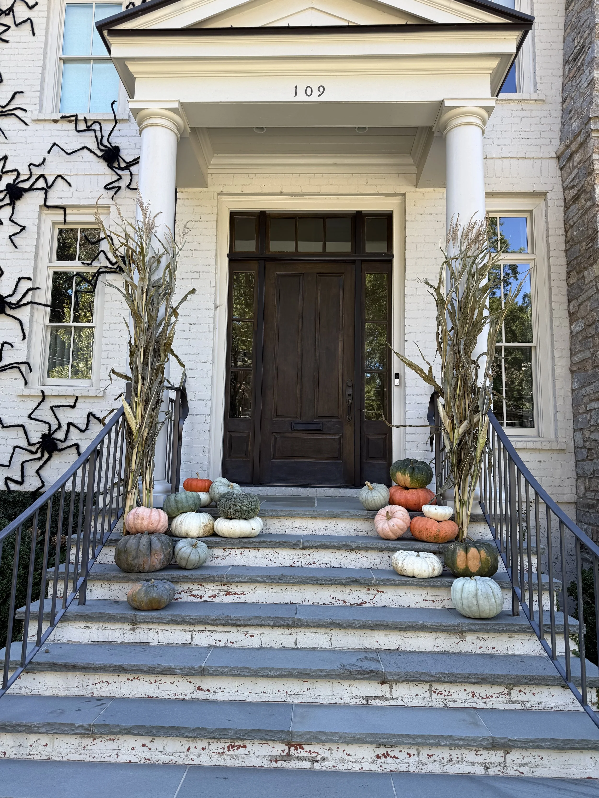 Front porch of a white brick house with black front door, decorated for fall with pumpkins, gourds, and dried corn stalks on the steps.