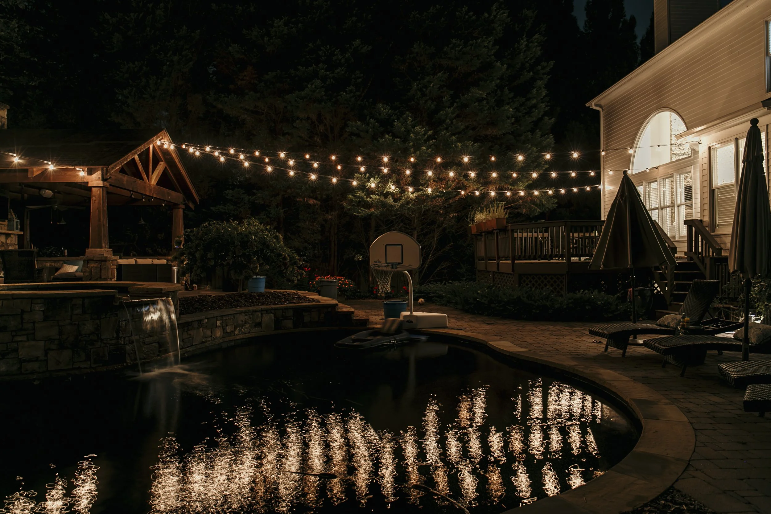 Night view of a backyard with a pool, string lights overhead, a small basketball hoop, lounge chairs, an umbrella, a deck with windows, and trees in the background. Light Up Atlanta