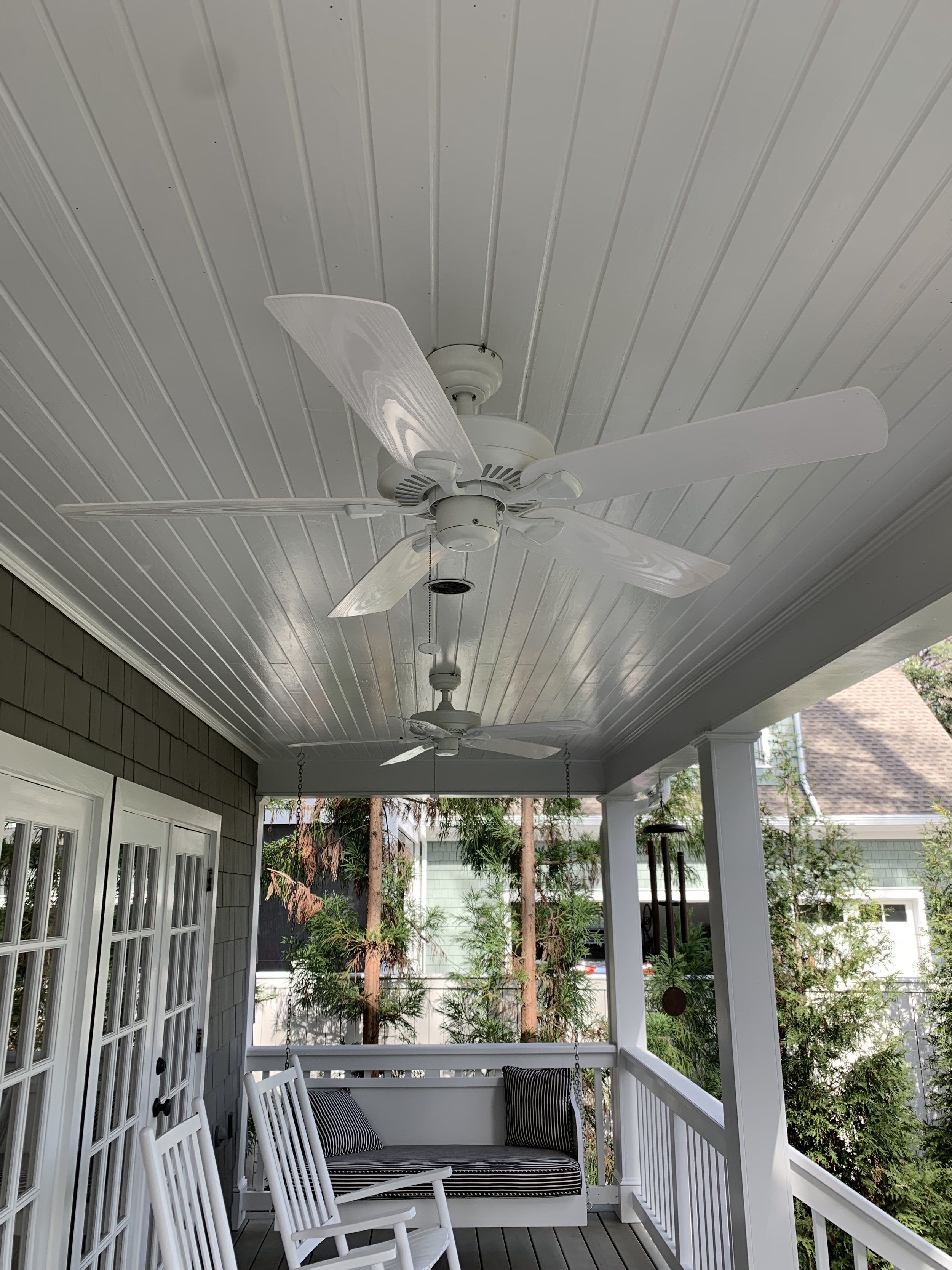 Covered porch with white ceiling fans, white rocking chairs, a porch swing with cushions, and greenery in the background.