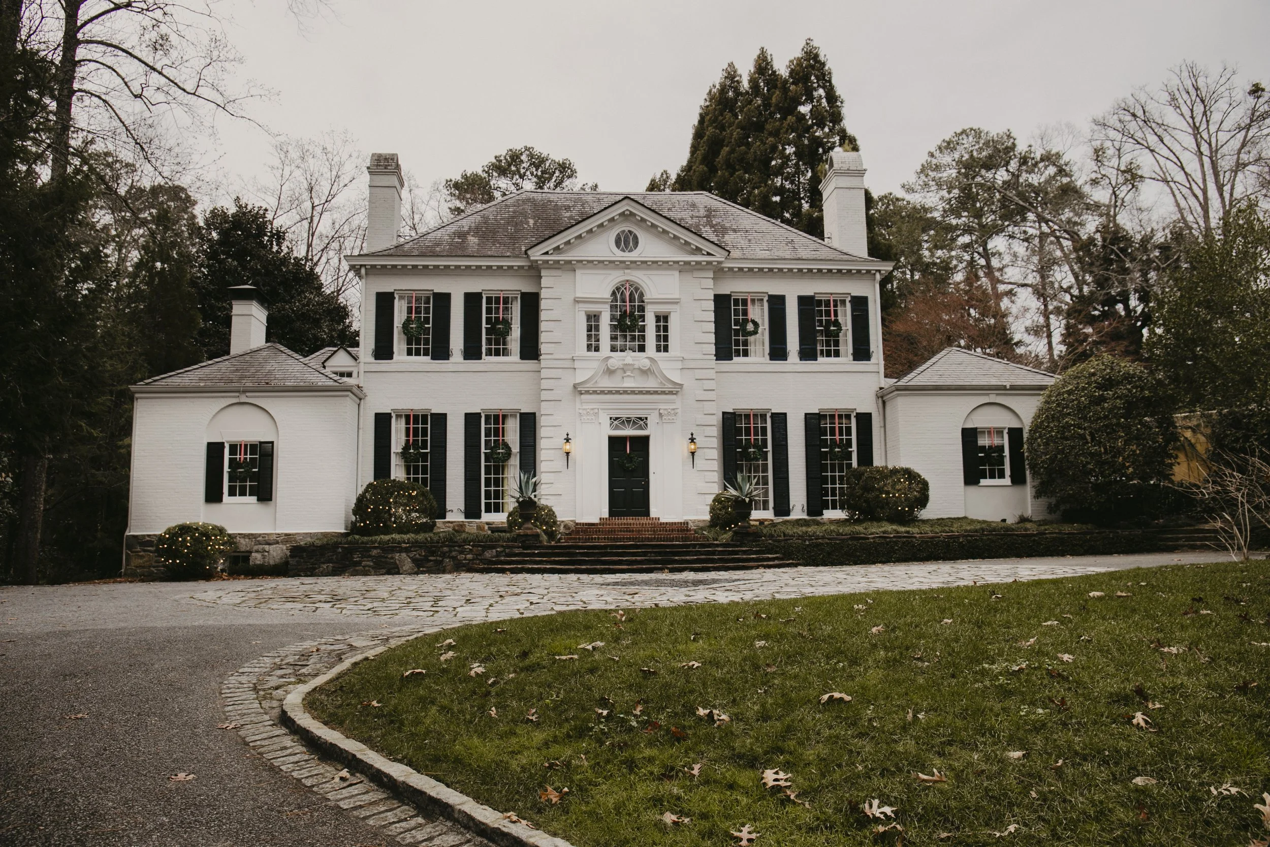 A large white mansion with black shutters, surrounded by leafless trees, with wreaths on the windows and bushes decorated with Christmas lights, and a driveway leading up to the front steps.