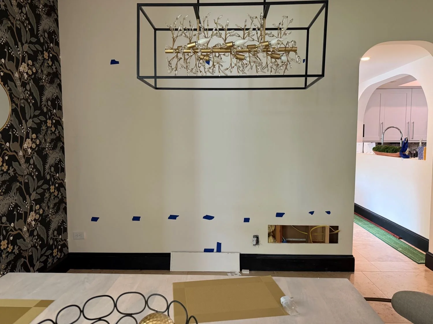 A dining room wall with taping for painting or renovation, showing blue painter's tape, an electrical outlet, and a cutout for wiring. The room has a decorative black and gold-themed wallpaper on the left, a modern chandelier hanging from the ceiling, and an open doorway revealing a kitchen with pink cabinets and a sink.