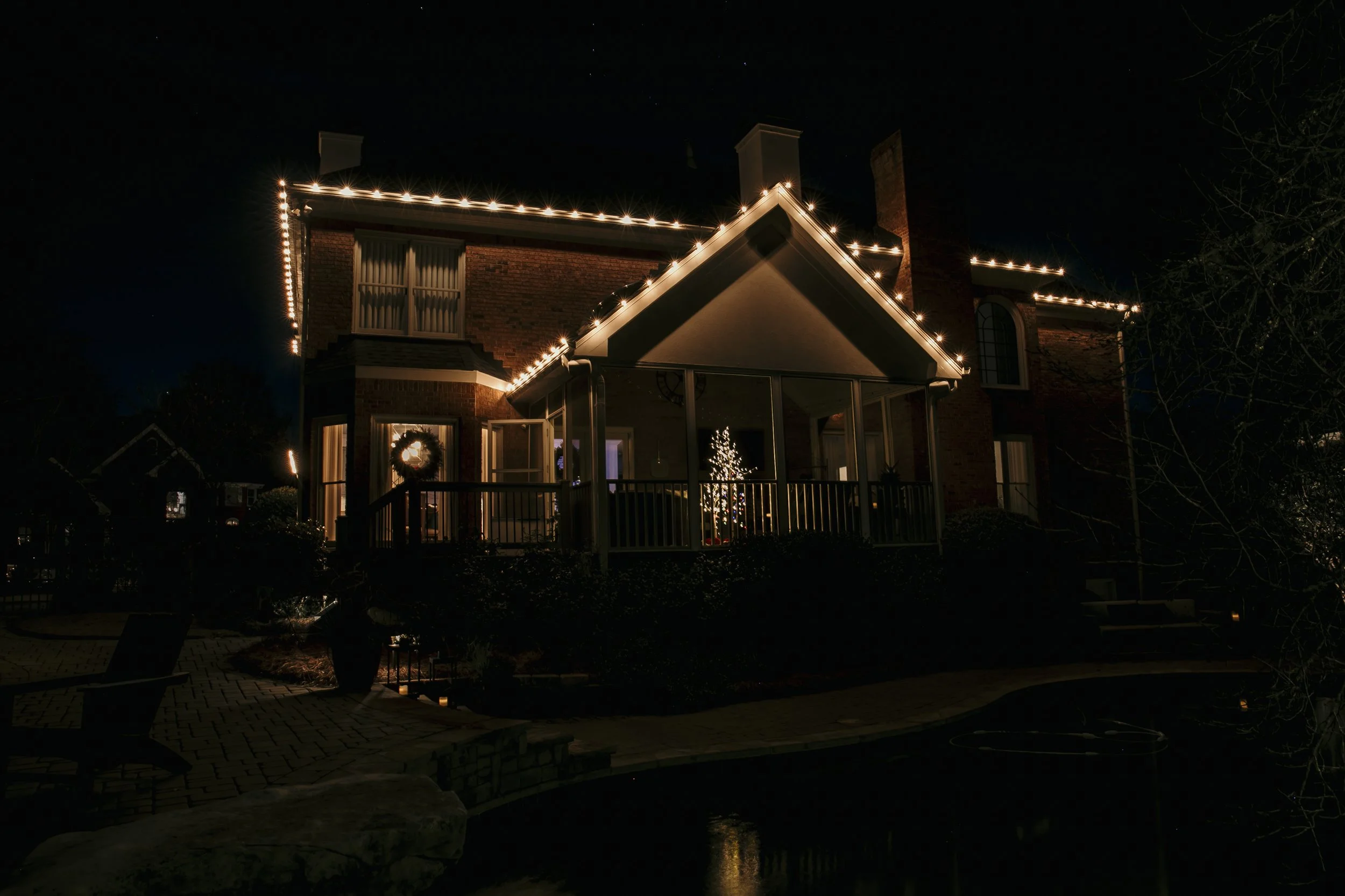 A brick house decorated with Christmas lights around the roofline and windows, with a lit Christmas tree visible through the large window on the porch at night.