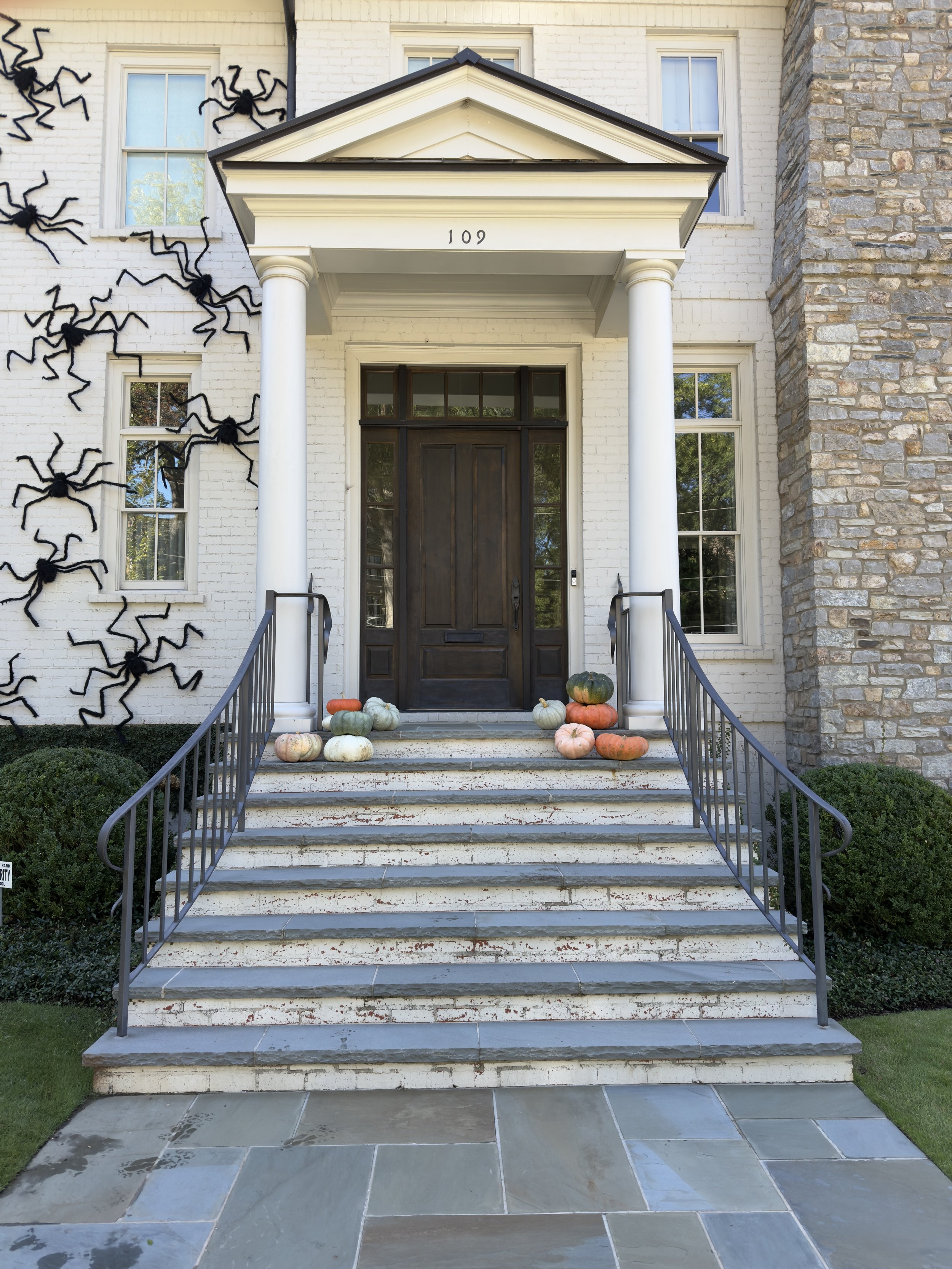 Front entrance of a house with white brick walls and stone accents, featuring a dark wooden door, white columns, and decorated with pumpkins and gourds, with Halloween spider decorations on the left side of the wall. Light Up Atlanta