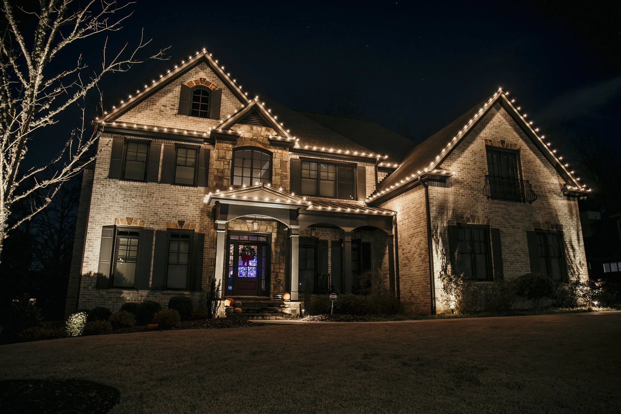A large brick house decorated with string lights outlining the roof and windows at night, with some shrubbery and a leafless tree in the front yard.