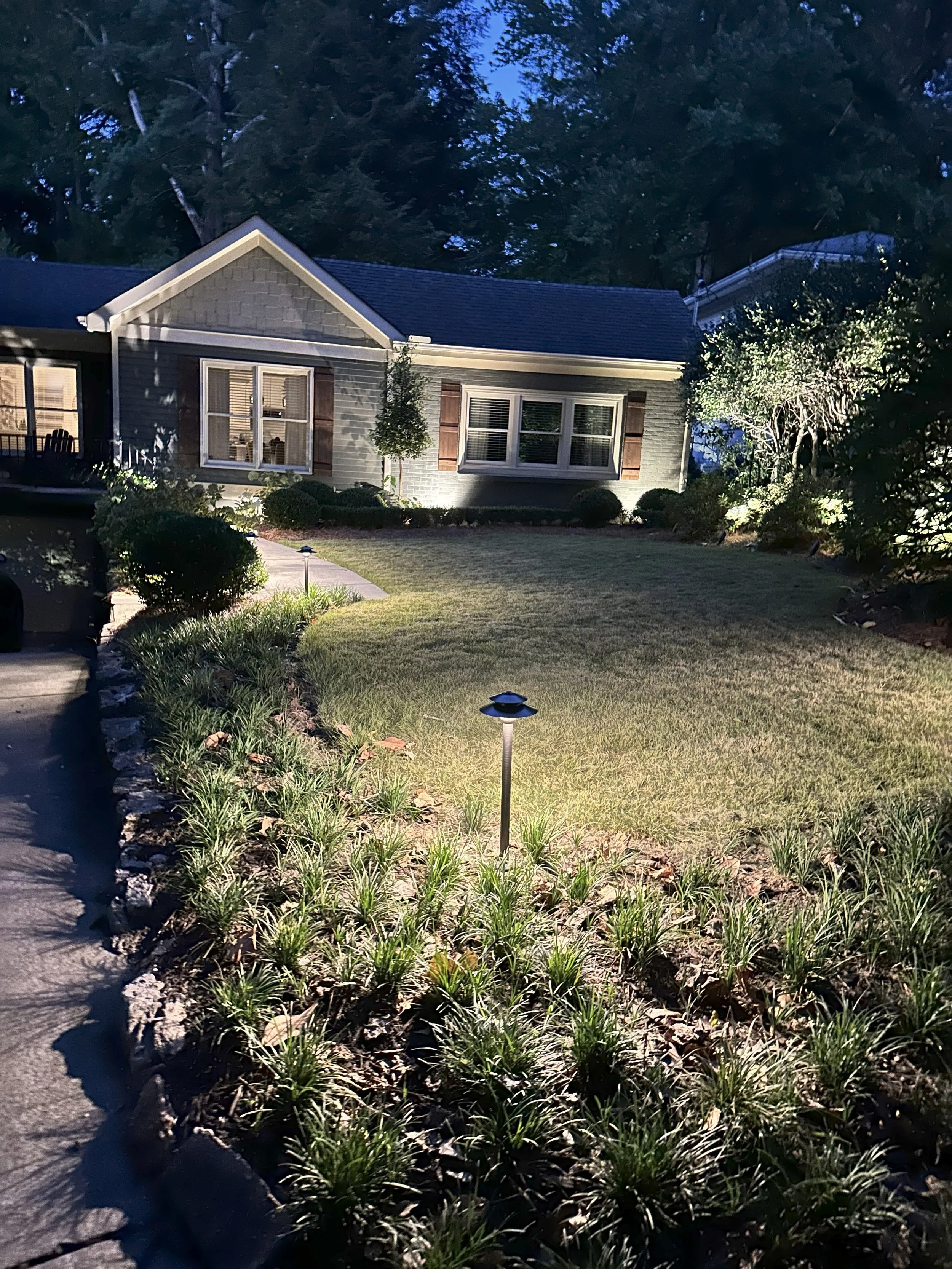Nighttime view of a well-lit front yard with a lawn, flower bed, and pathway leading to a house with large windows and dark shutters, surrounded by trees.