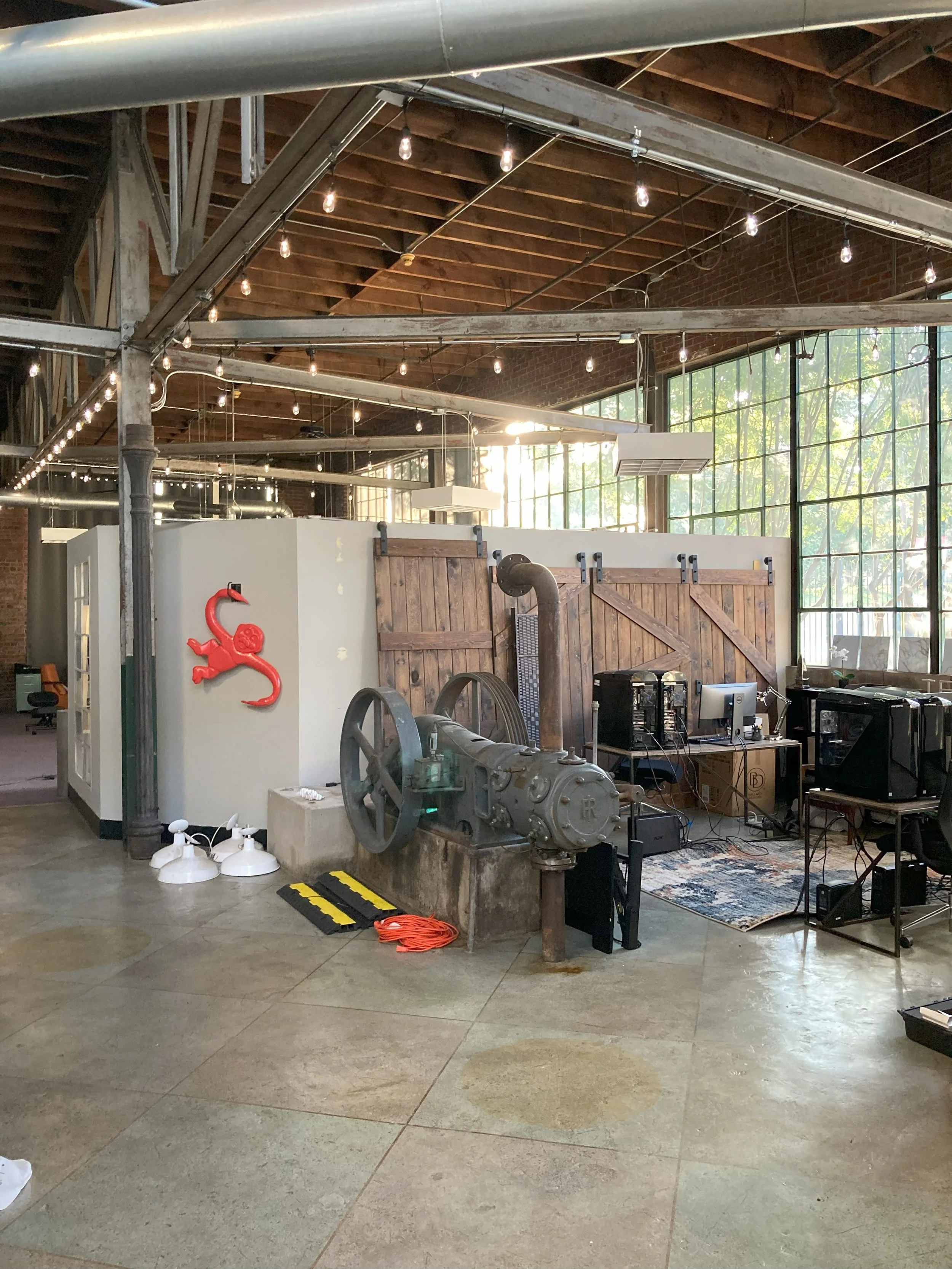 Interior of a renovated industrial-style office space with large windows, exposed brick and wood ceiling, string lights, vintage machinery, computers, and a small red wall decoration of a gecko. Light Up Atlanta