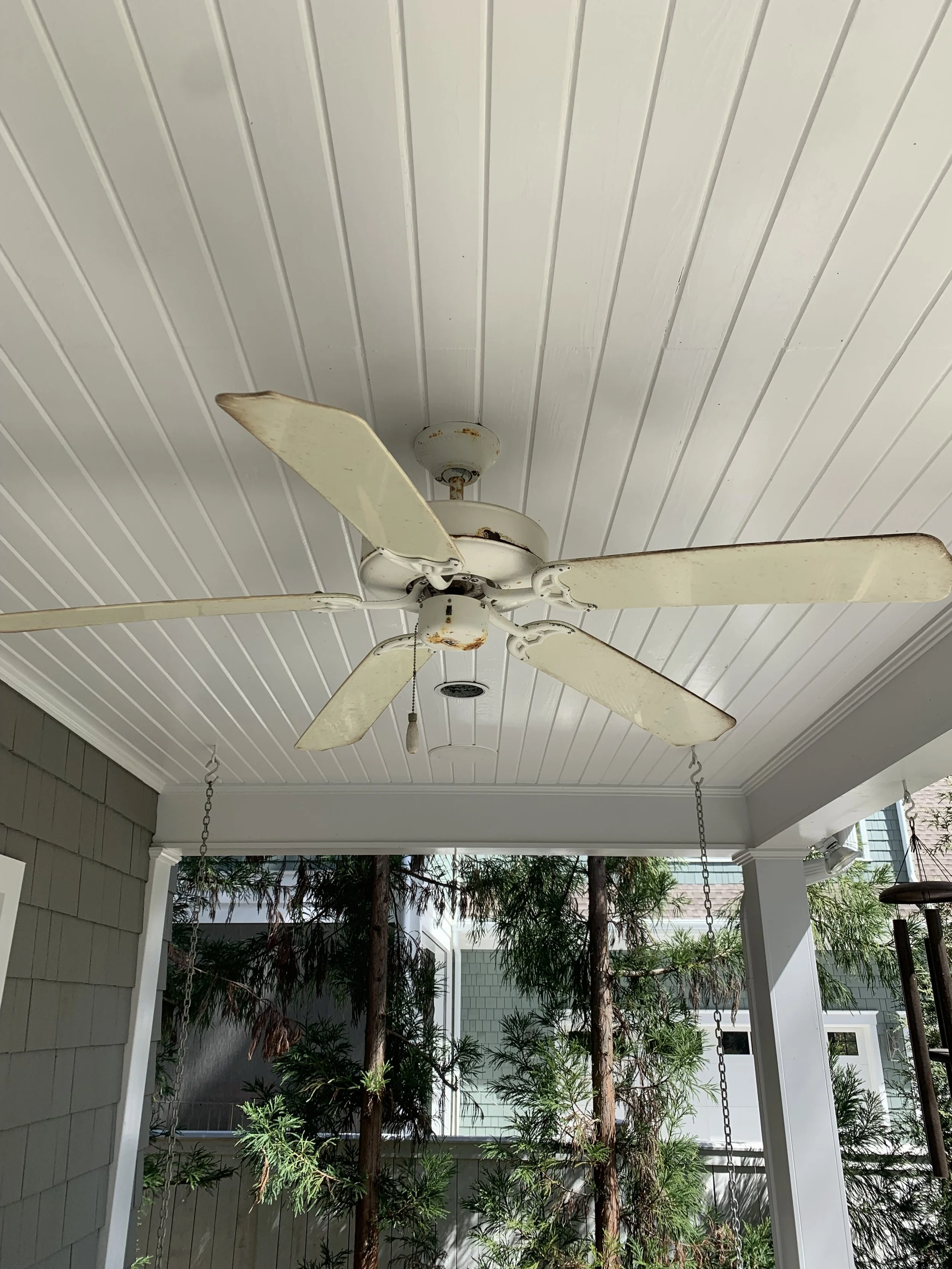 An old, white ceiling fan with rust spots, hanging from a white beadboard ceiling on a porch, with chains holding the fan, and green trees visible outside. Light Up Atlanta