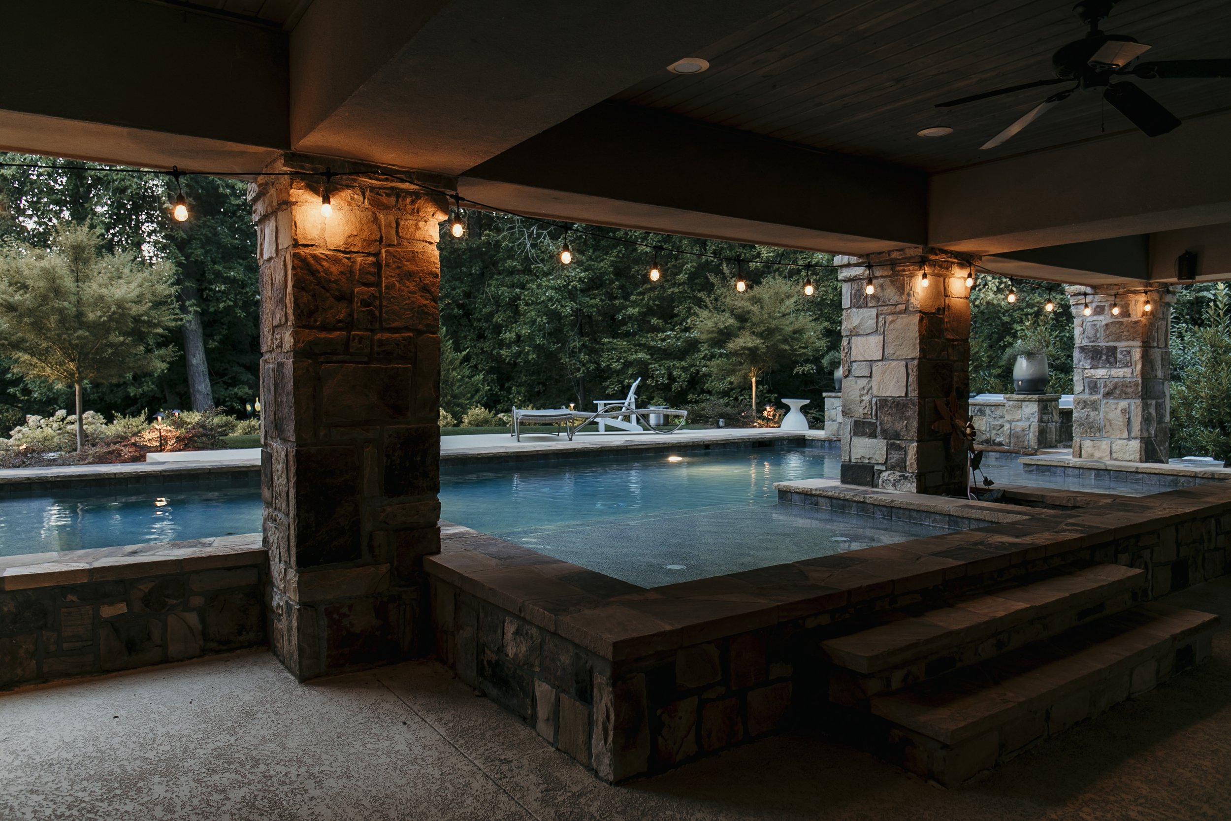 Undercover patio with a view of a swimming pool, stone columns, string lights, outdoor chairs, and lush green garden in the evening. Light Up Atlanta