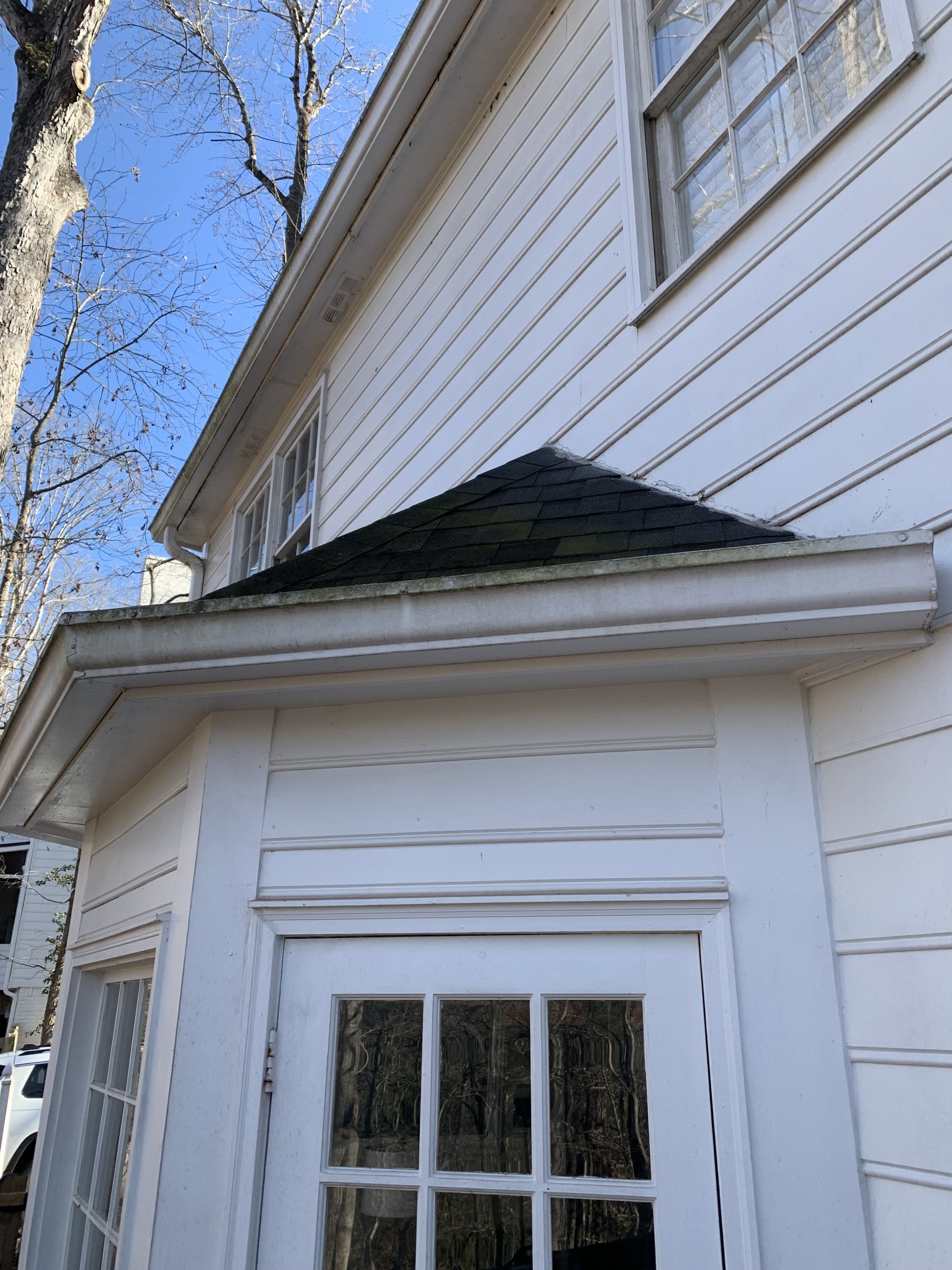 Close-up of the upper part of a white house with a sloped roof, gable, and window, surrounded by leafless trees under a clear blue sky. Light Up Atlanta