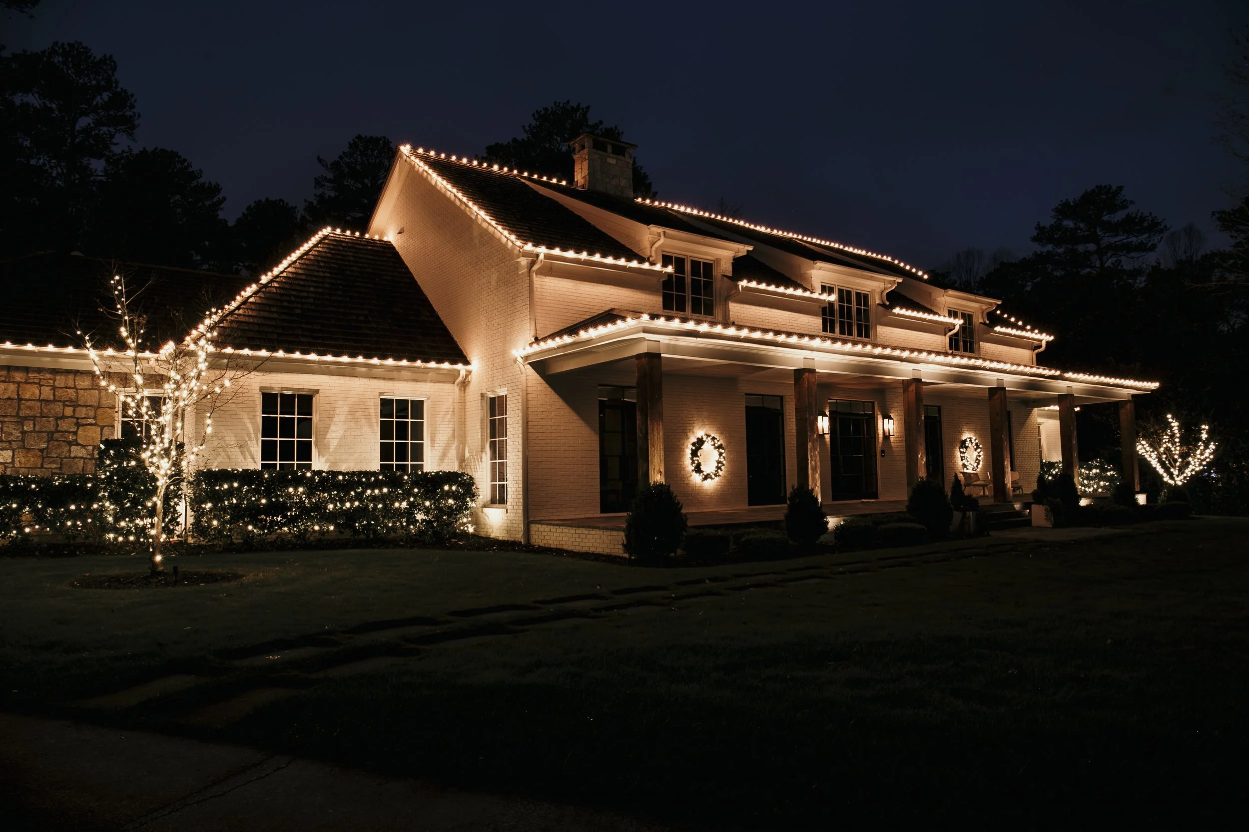 A large house decorated with white Christmas lights along the roof edges, windows, and bushes in front. Two illuminated wreaths are hung on the front wall, and there are lit trees on each side of the house. The house is at night against a dark sky.