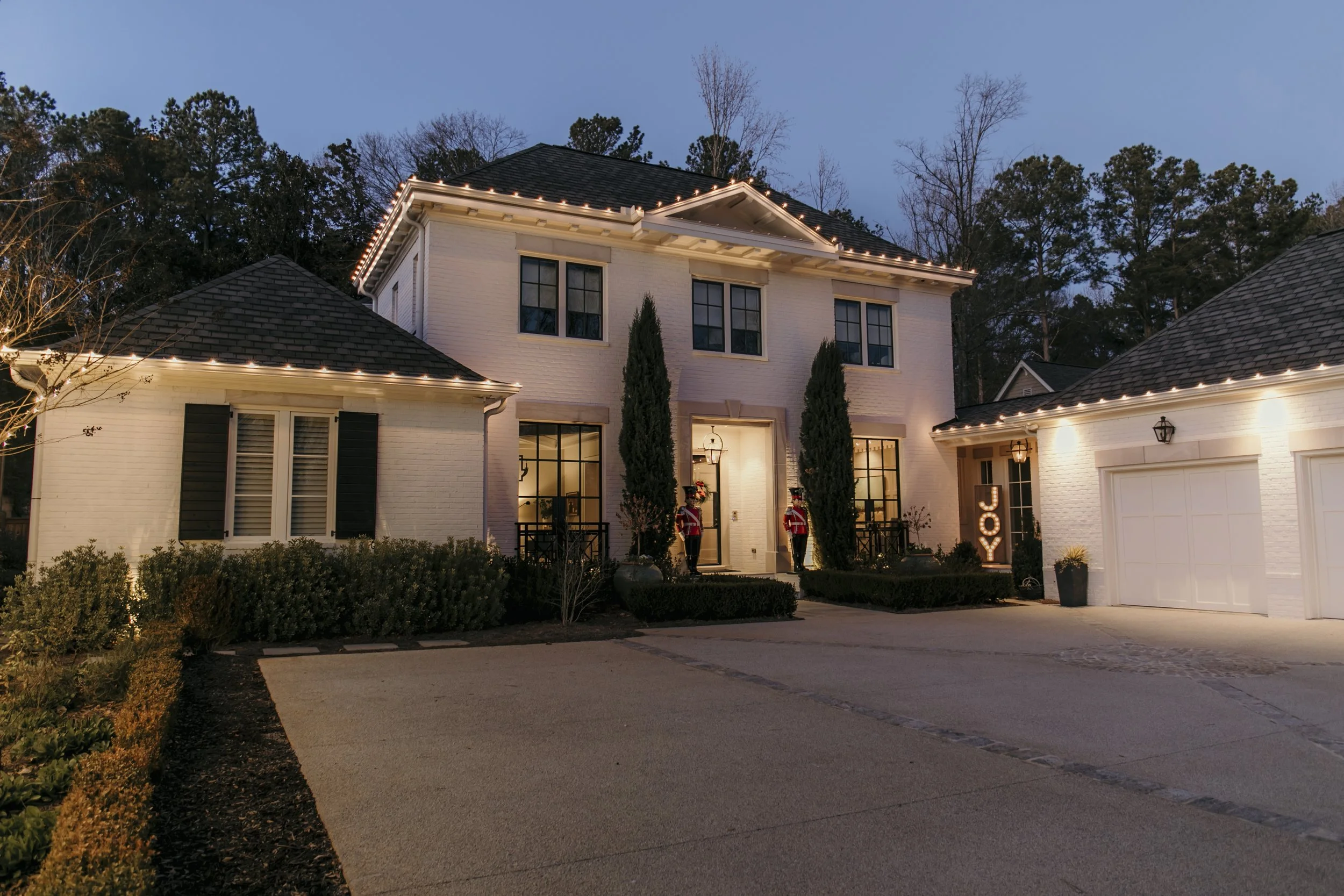 A large white two-story house decorated for Christmas with string lights along the roofline. There are two nutcracker statues flanking the front door, and a lit sign that reads "JOY" to the right of the entrance. The driveway is visible in the foreground with surrounding landscaped bushes and trees in the background.
