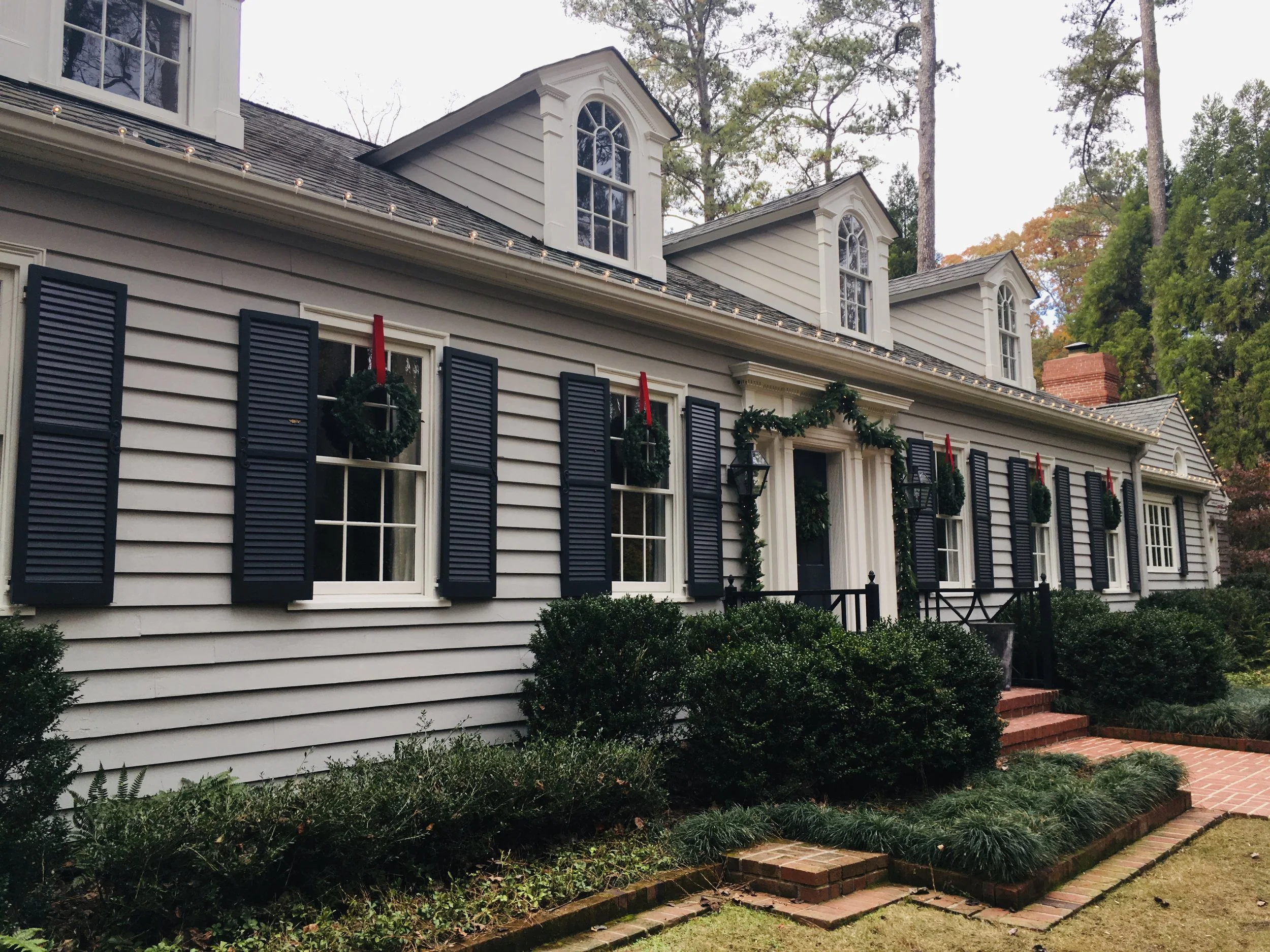Front view of a house decorated with Christmas wreaths on window shutters and garlands around the door, surrounded by greenery and trees. Light Up Atlanta