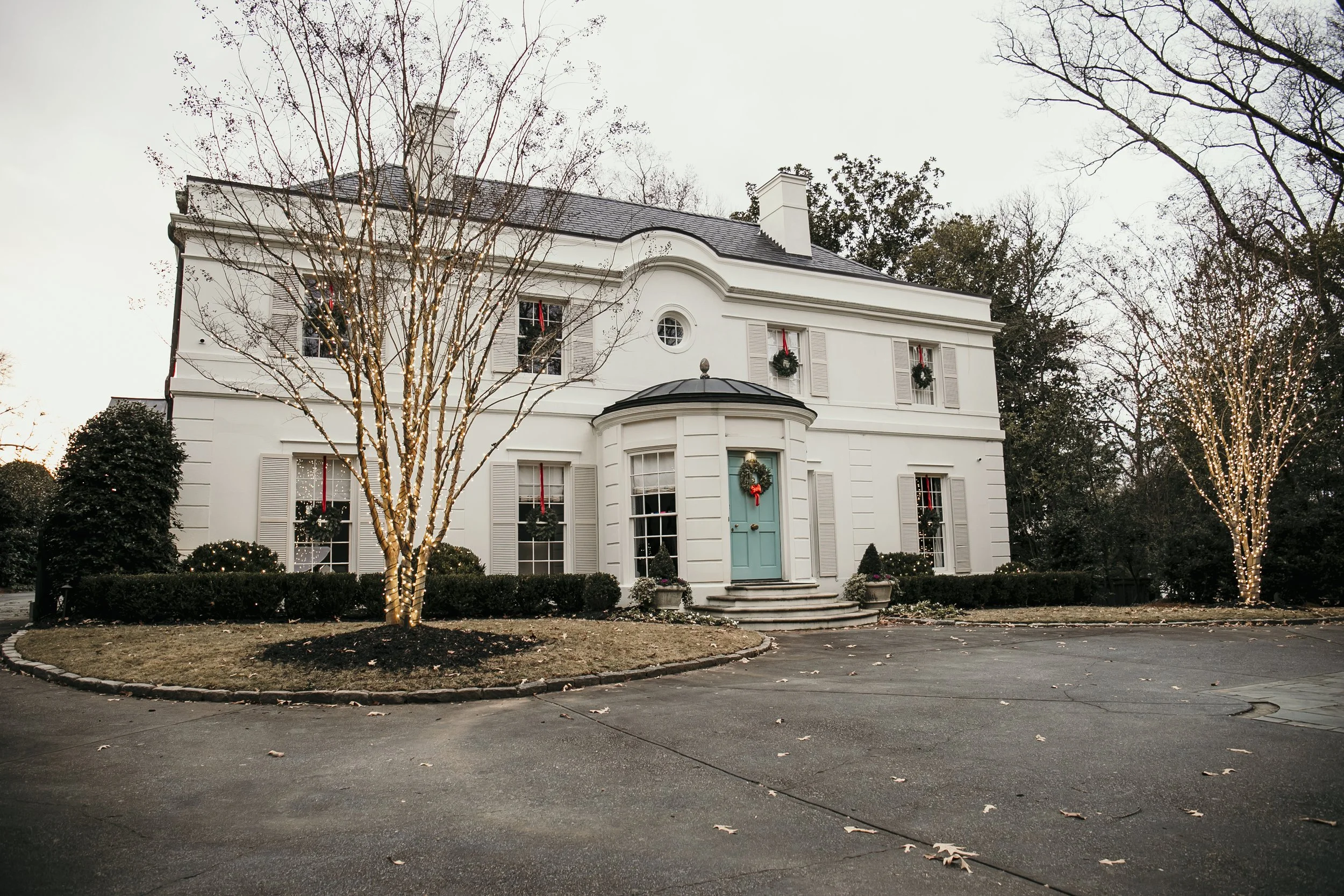 A large white house decorated with Christmas wreaths, standing in a driveway with leafless trees wrapped in holiday lights. Light Up Atlanta