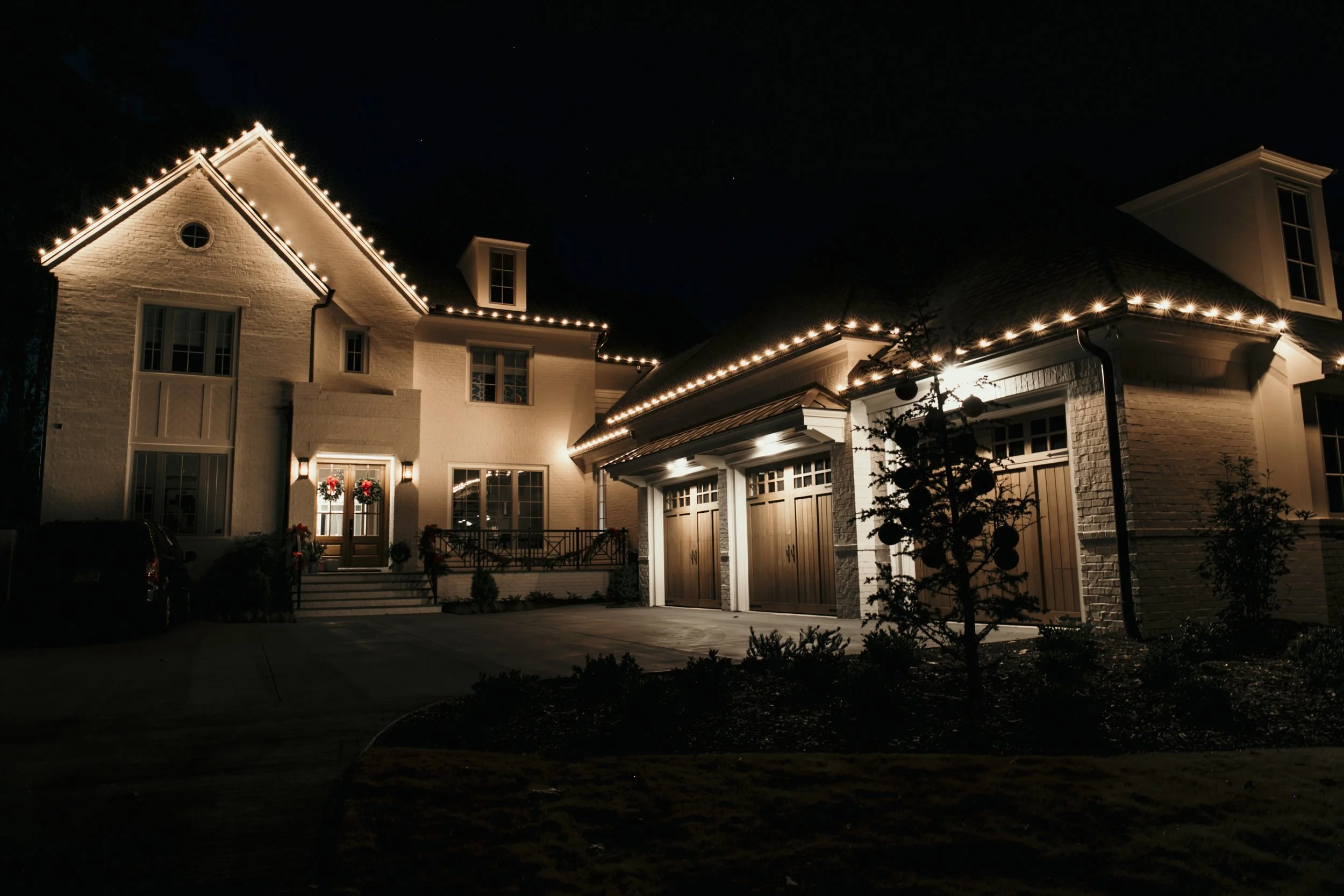 Nighttime view of a large house decorated with Christmas lights and wreaths, white exterior, with garage doors and front steps. Light Up Atlanta