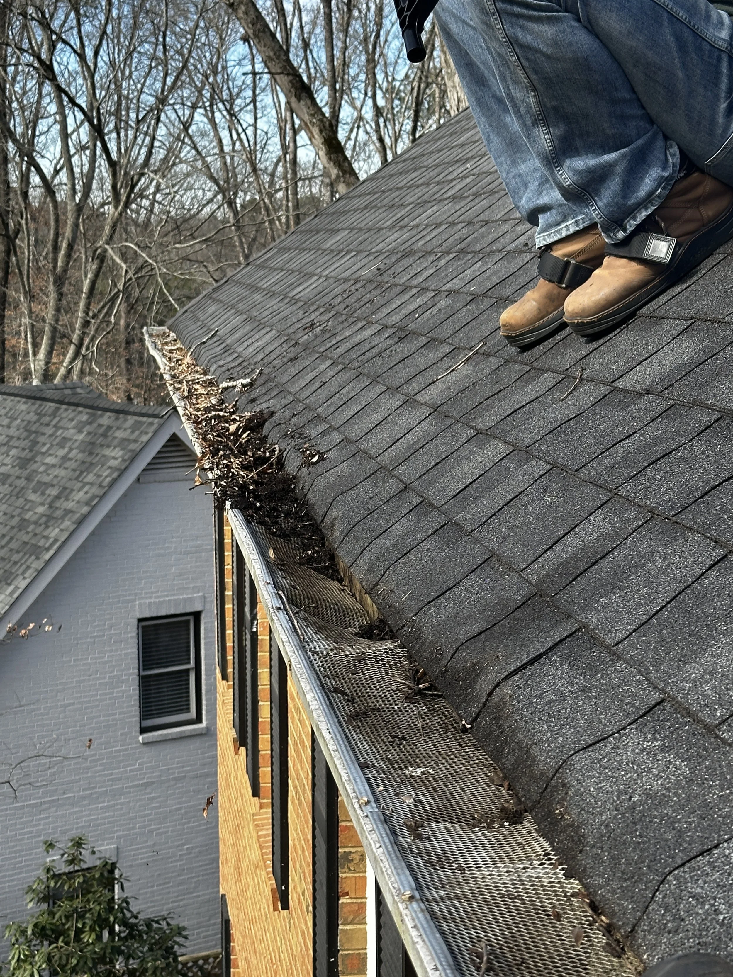 Person on a roof inspecting or cleaning the gutter, wearing work boots and jeans, with bare trees in the background. Light Up Atlanta
