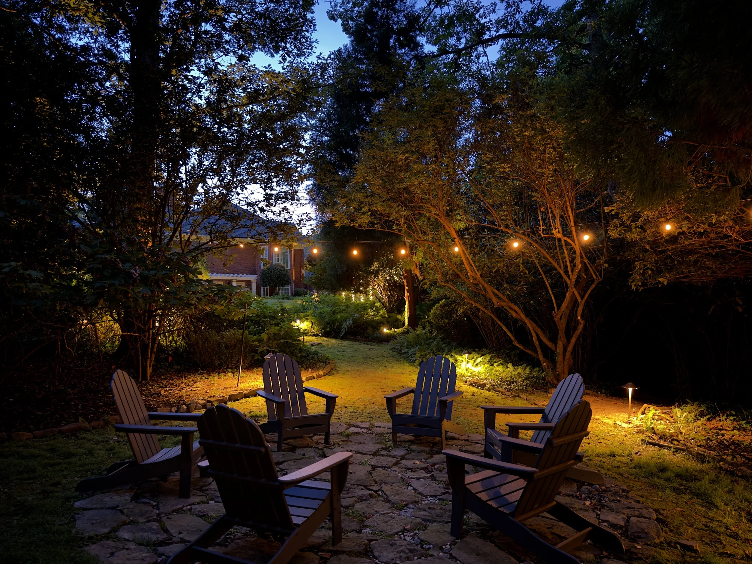 A backyard area with five Adirondack chairs arranged in a circle on a stone-paved patio, surrounded by trees, shrubs, and garden lighting, with a house in the background during dusk. Light Up Atlanta
