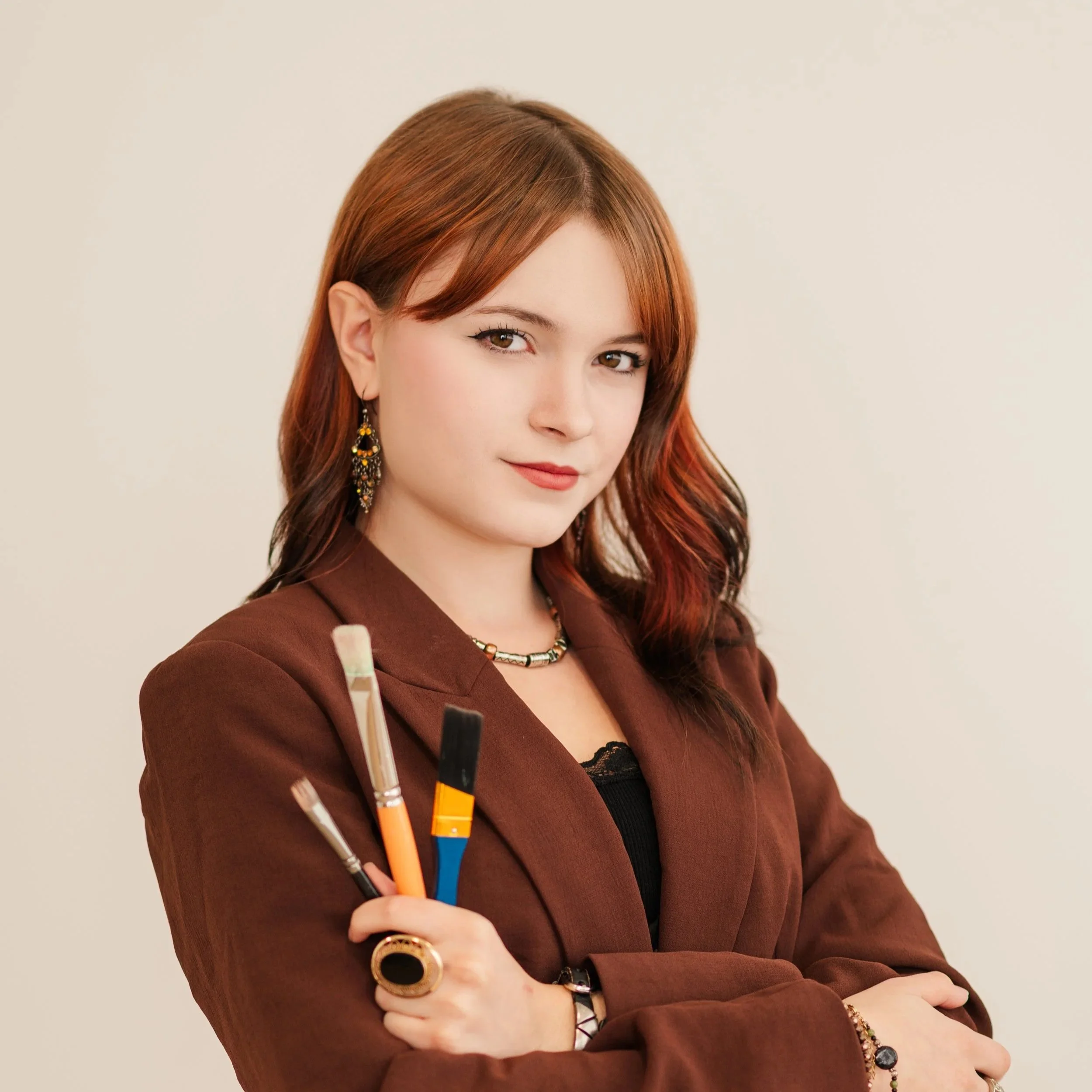 A woman with brown and red hair holding paintbrushes, wearing a brown blazer, jewelry, and looking confidently at the camera.