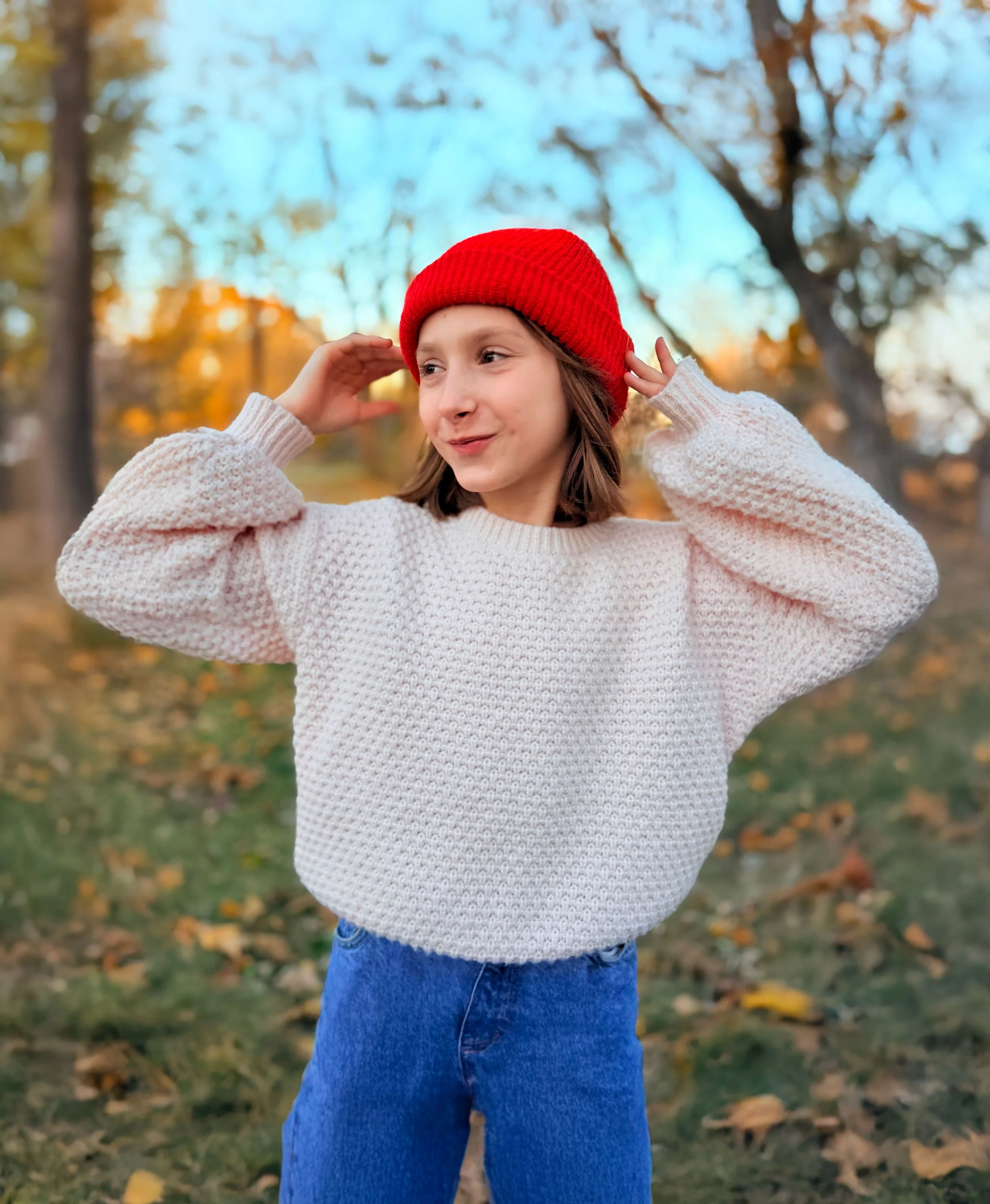 A young girl outdoors in autumn, wearing a red knit hat, a cream-colored sweater, and blue jeans, with trees and fallen leaves in the background.
