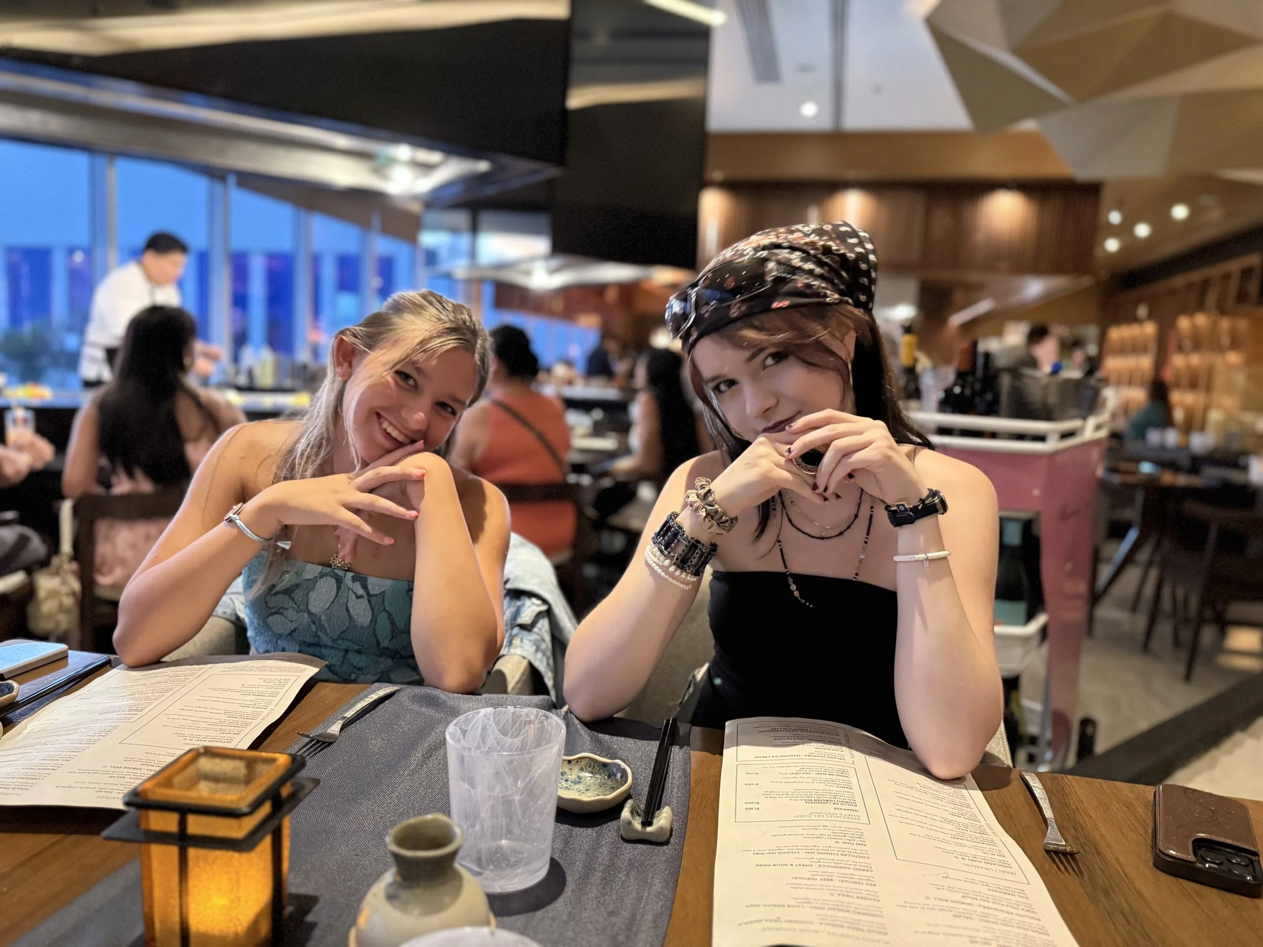 Two young women sitting at a restaurant table, smiling and posing for the photo, with menus, a glass, and small dishes on the table. The background shows other diners and a busy restaurant interior.