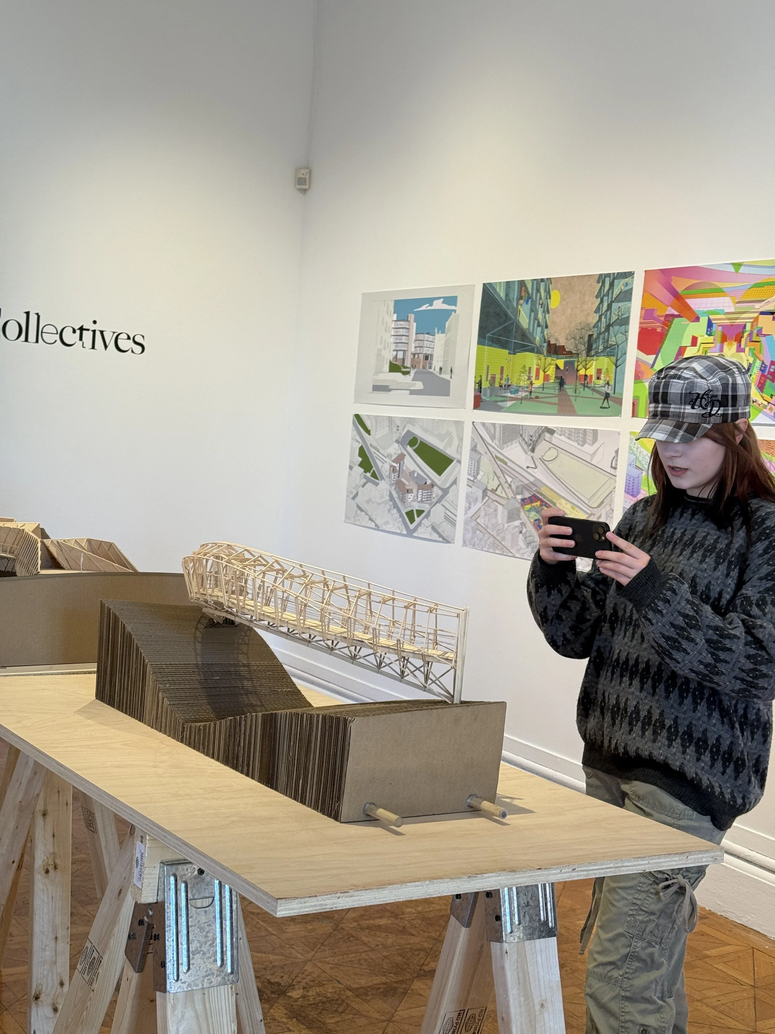 A person wearing a plaid cap and a dark sweater is using a smartphone in a gallery or exhibition space. They are looking at a wooden architectural model on a table. Behind them on the wall are colorful architectural renderings or plans.