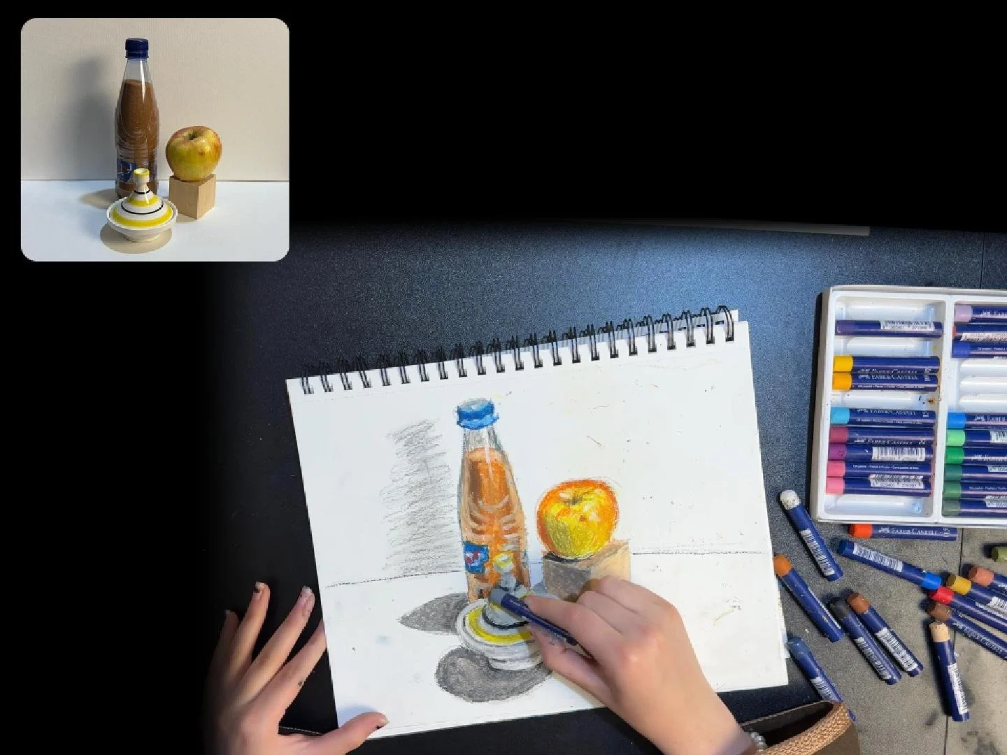 A child’s drawing of a plastic bottle, an apple on a stand, and a pencil sketch, with oil pastels and crayons scattered around on a desk.