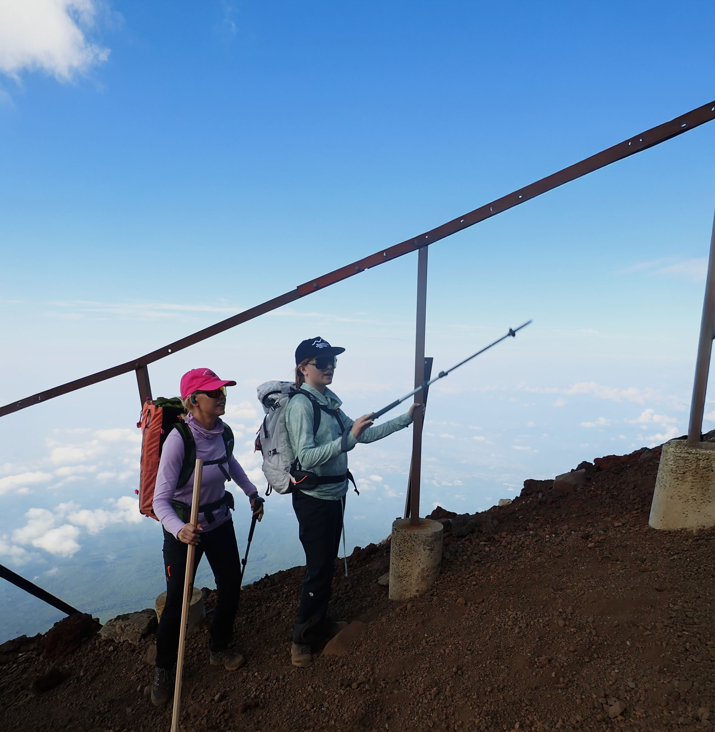 Two women on a mountain trail, wearing hiking gear, with one woman pointing a trekking pole toward a protective railing overlooking clouds and sky.