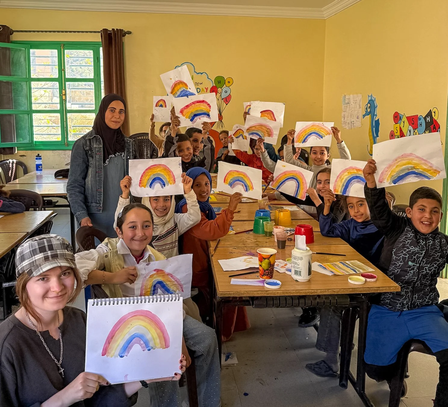 Group of children and a woman in a classroom holding up drawings of rainbows.