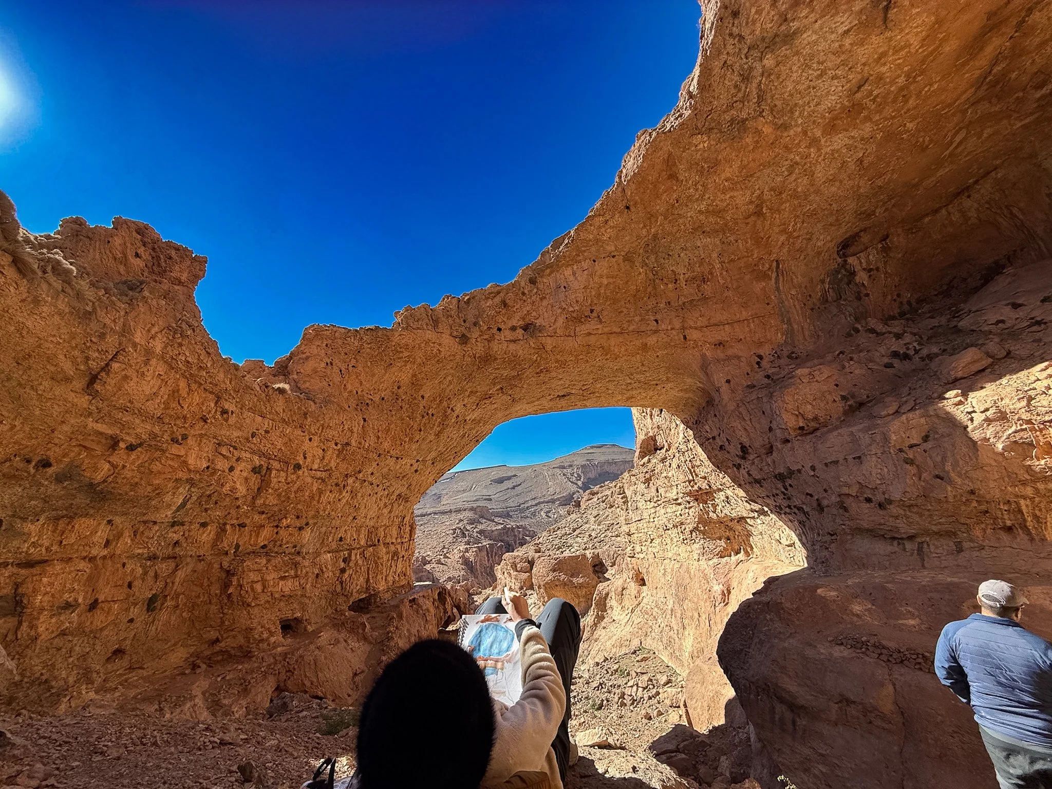 People sitting and relaxing inside a natural rock formation or cave with large openings, overlooking a desert landscape under a clear blue sky.