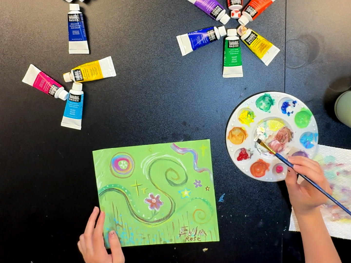 Art supplies including paint, paintbrushes, and colorful artwork on paper are arranged on a table, indicating an art activity.