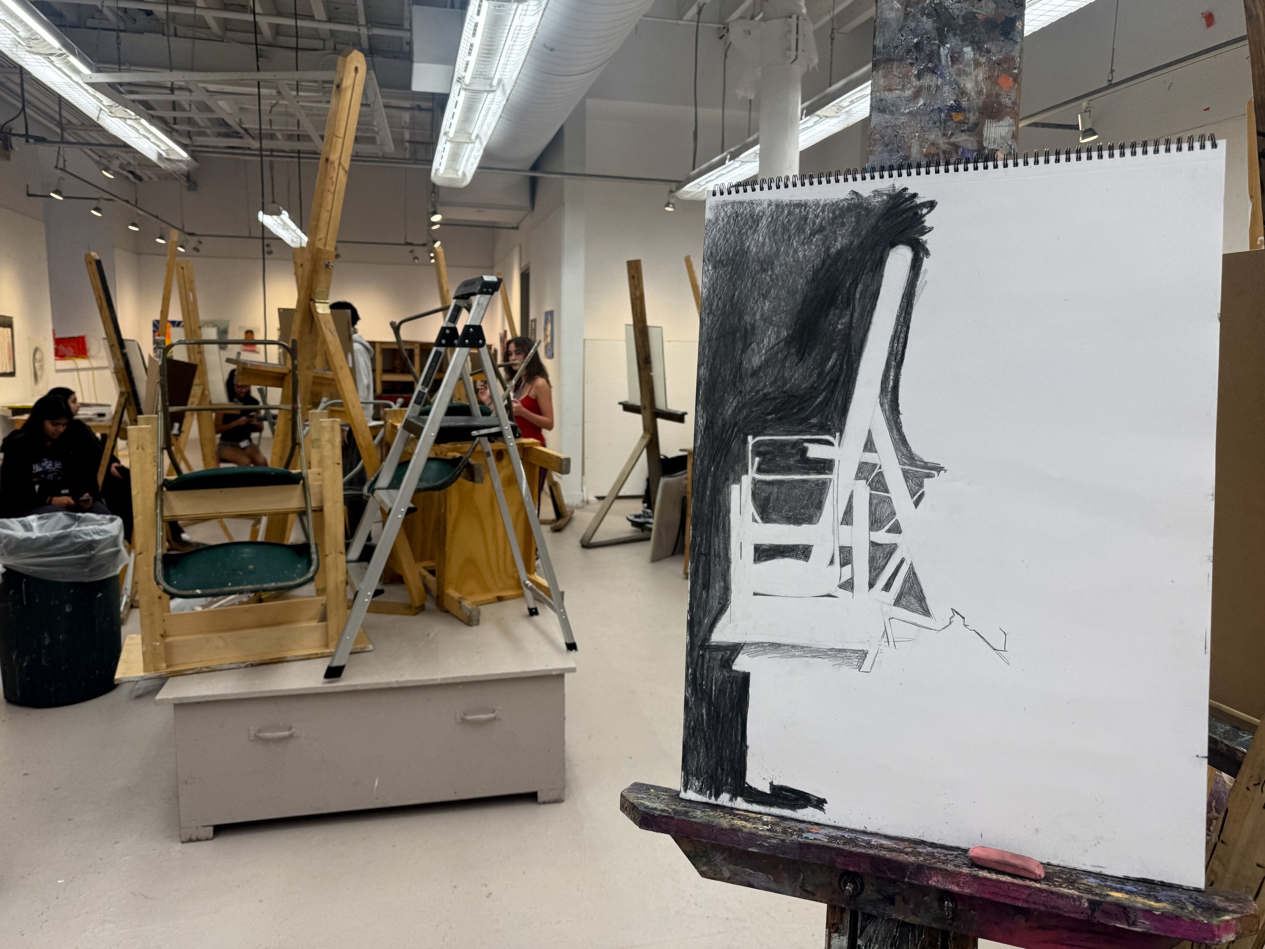 An art gallery or studio with people working on paintings, wooden easels, a ladder, and a large black and white painting of a chair and a table visible in the foreground.
