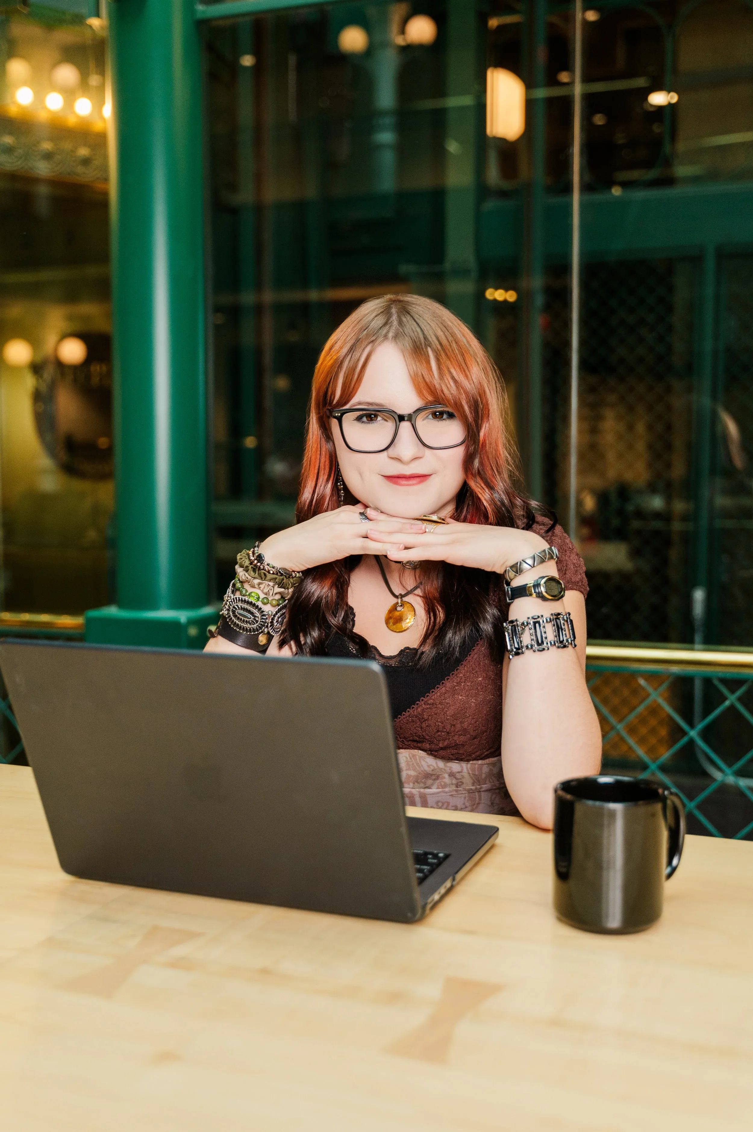 A woman with red hair and glasses sitting at a desk with a laptop, a black coffee mug, and wearing multiple bracelets and necklaces.